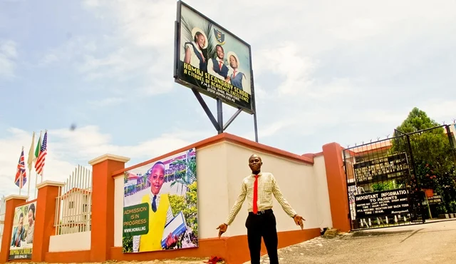 A man standing outside a school gate with two large digital screens displaying school information and advertisements, and a billboard above showing a TV show or movie poster. The school gate has flags and an orange and beige wall, with trees and a cl