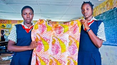 Two schoolgirls in uniform holding up a colorful fabric in a classroom.