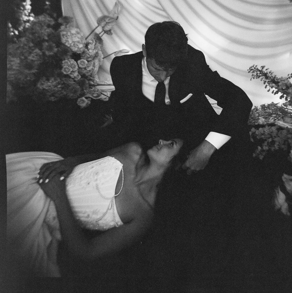 A man in a black suit and a woman in a white dress indoors with floral decorations, with the woman lying on the floor and the man leaning over her, looking at her.