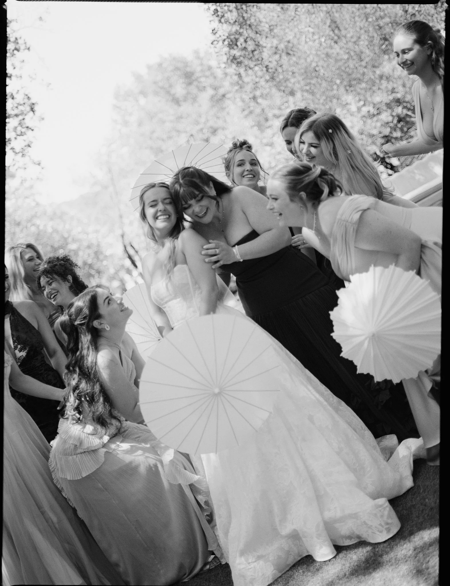 Group of women in formal dresses gathered outdoors, smiling and laughing, holding parasols on a sunny day.