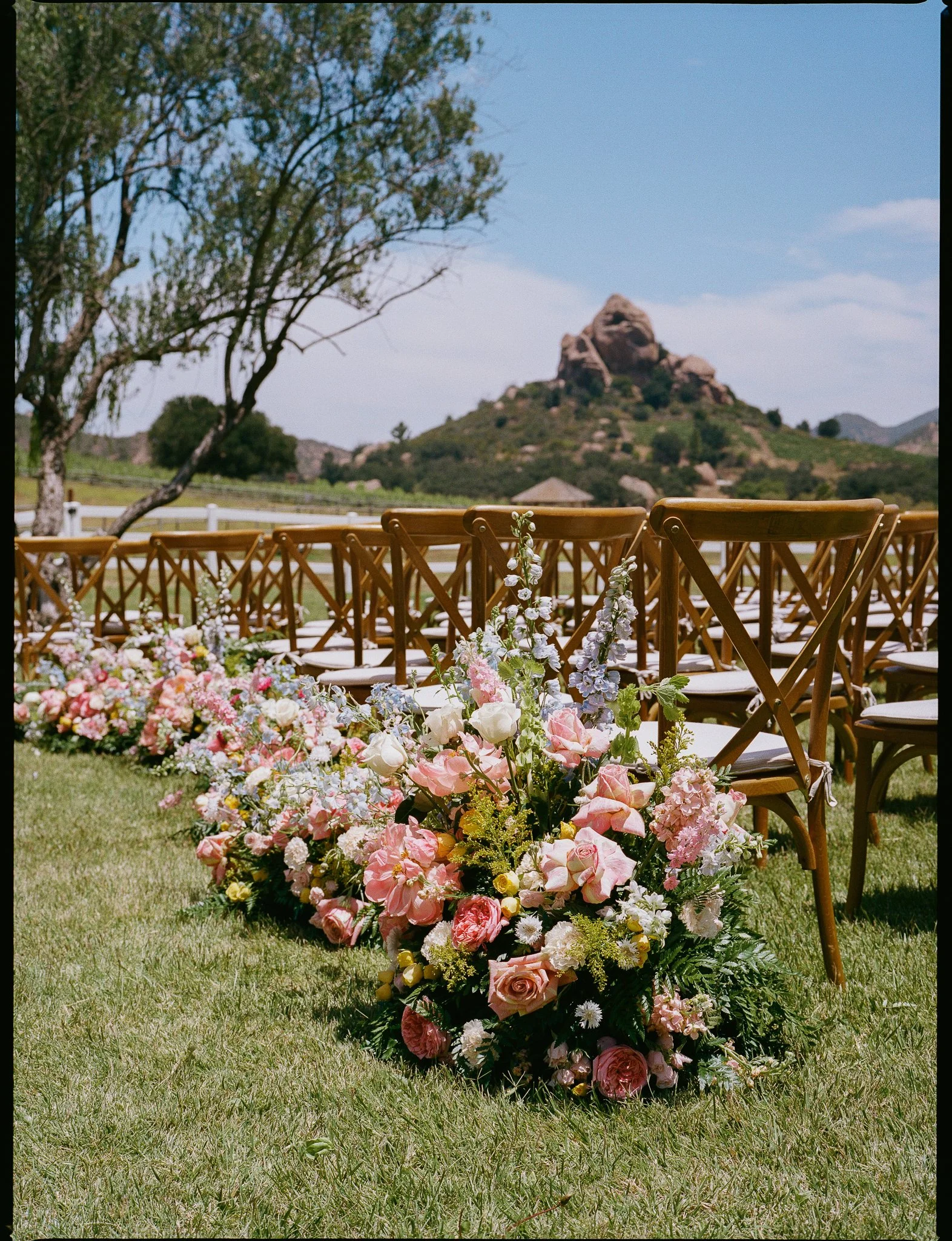 A large floral arrangement with pink, white, and yellow flowers and greenery on a grassy outdoor area, with wooden chairs and a scenic mountain with a prominent rock formation in the background under a clear blue sky.