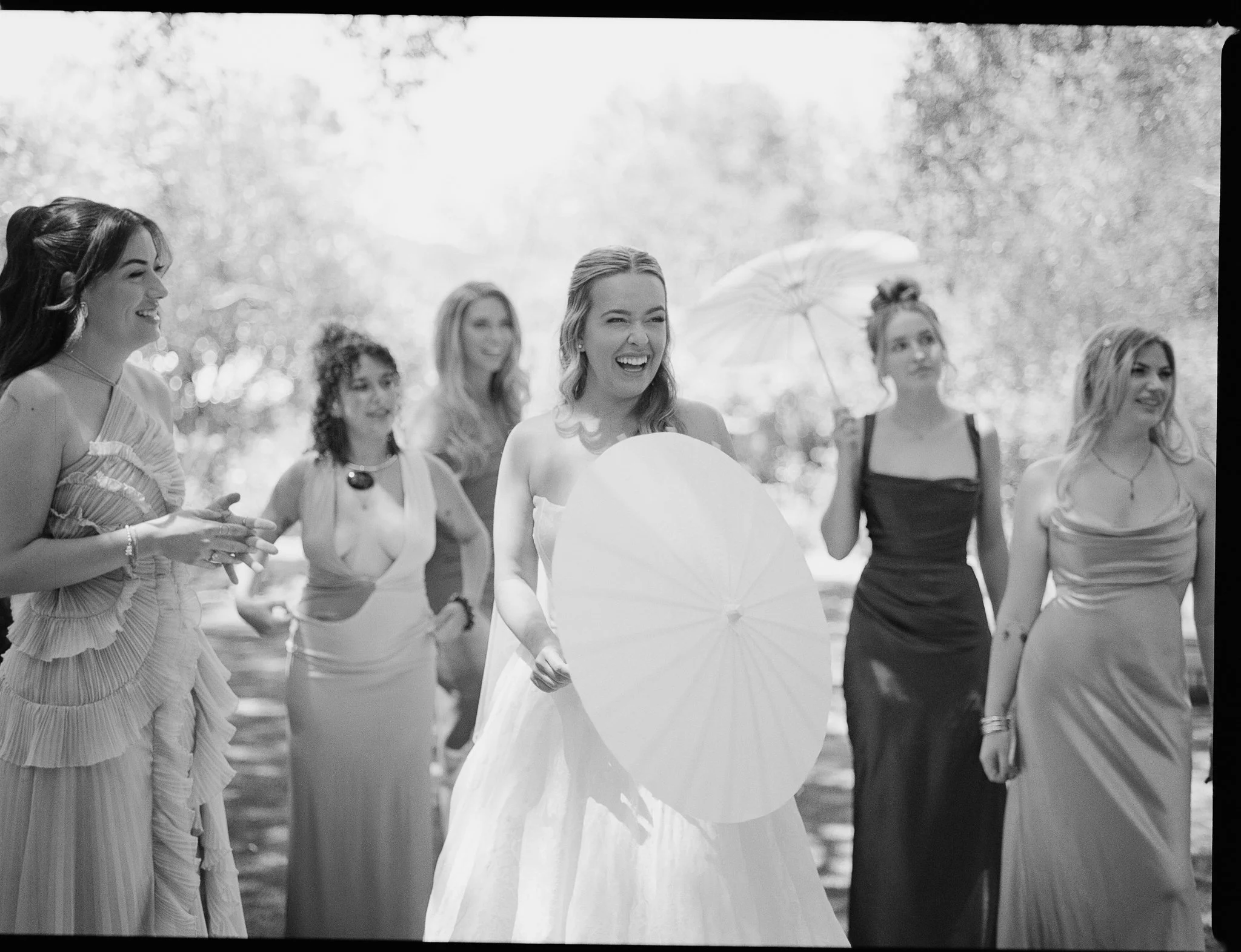 Group of women dressed in formal attire, outdoors, with one woman holding a parasol, smiling and laughing.