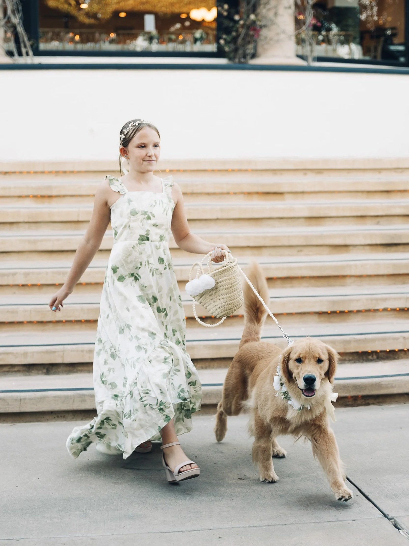 A young girl in a floral dress walks down steps with a golden retriever, both wearing floral accessories, during an outdoor event at sunset.