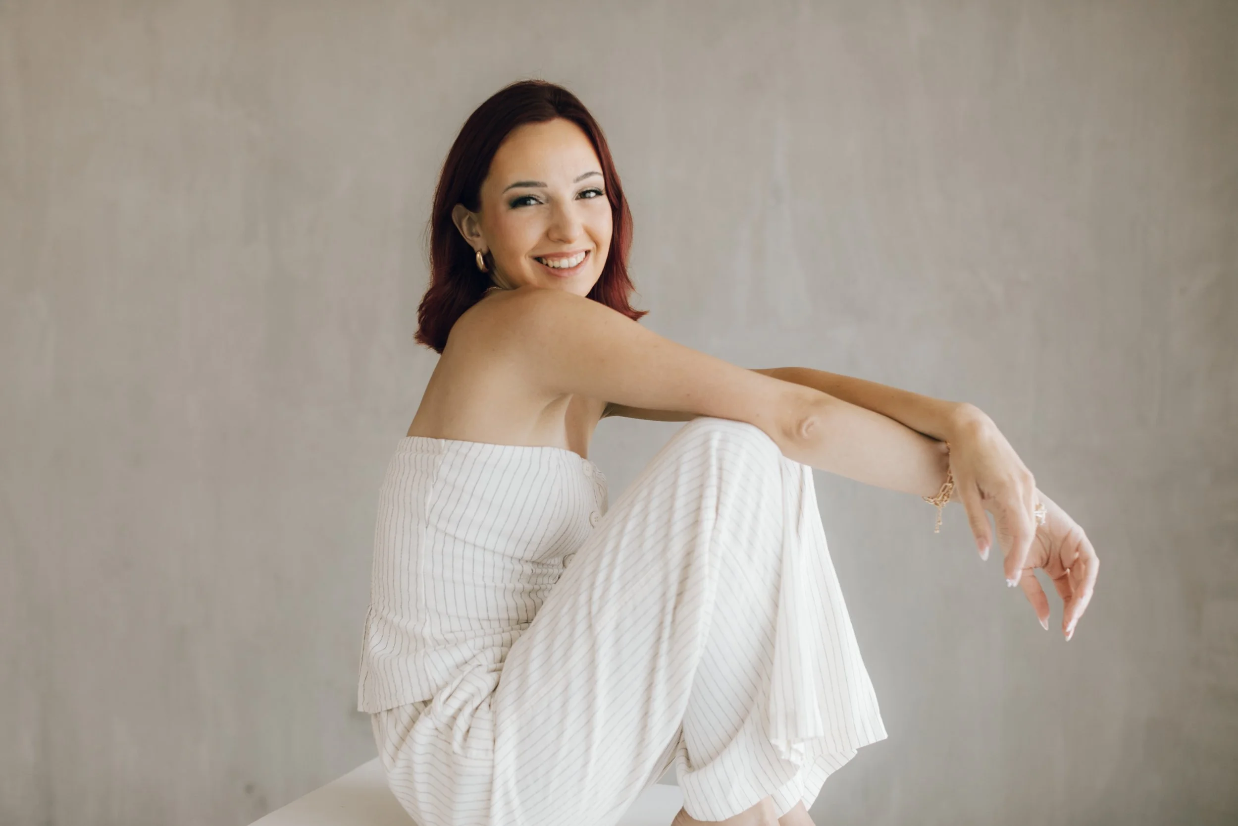 A woman with red hair, wearing earrings and a white striped off-shoulder dress, smiling and sitting with her arms wrapped around her knees against a plain wall.