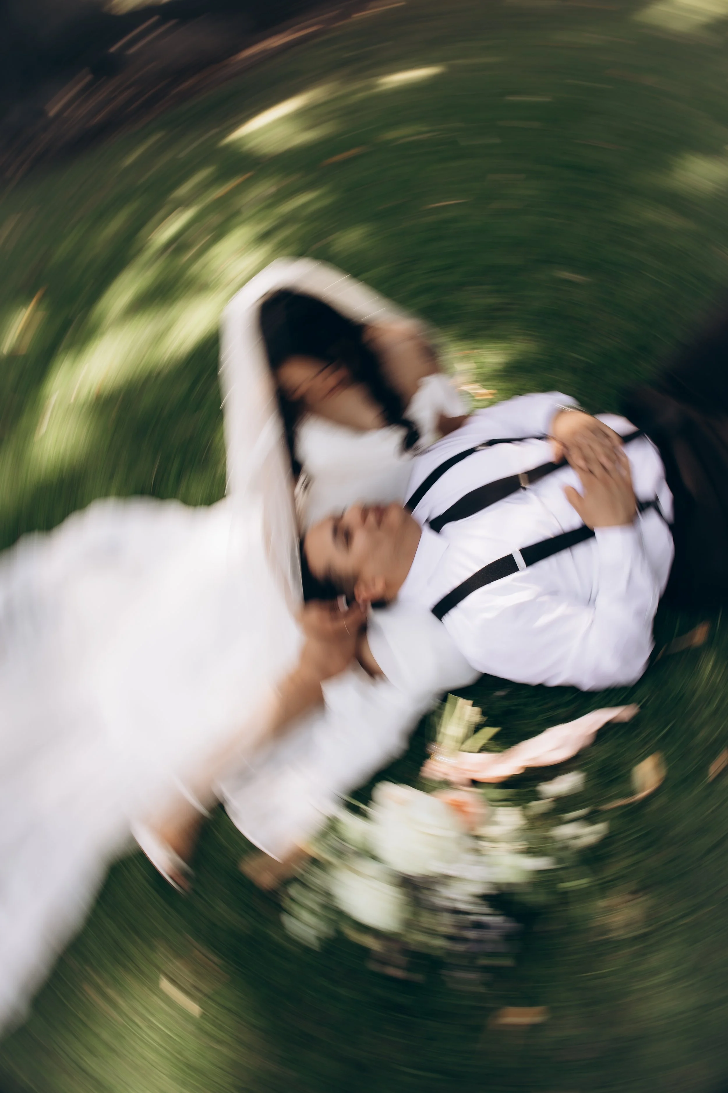 A woman and a man dressed in wedding attire, embracing on a lawn with a blurry, circular motion effect in the background.