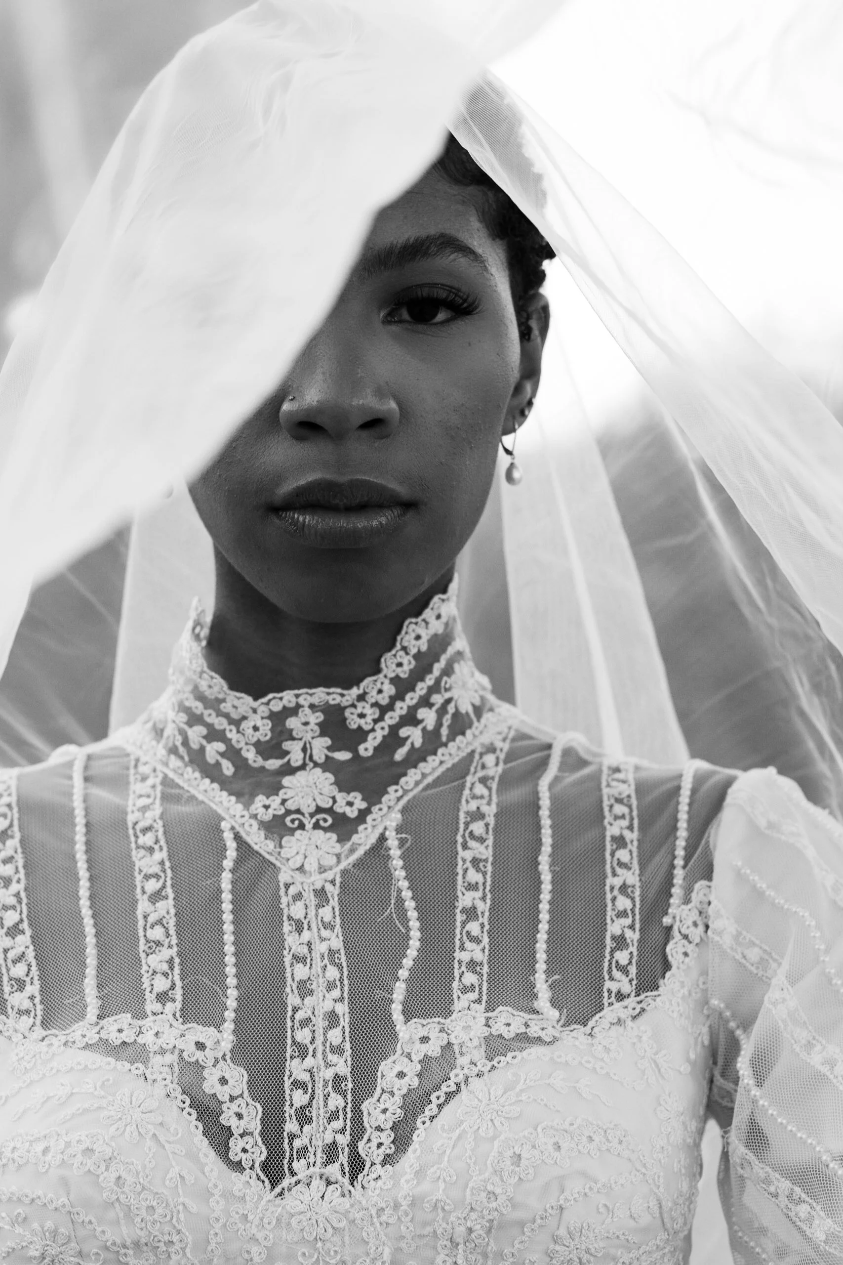 Close-up black and white photo of a woman partially covered by a light, sheer veil, showcasing her eye, nose, lips, earring, and detailed lace dress.