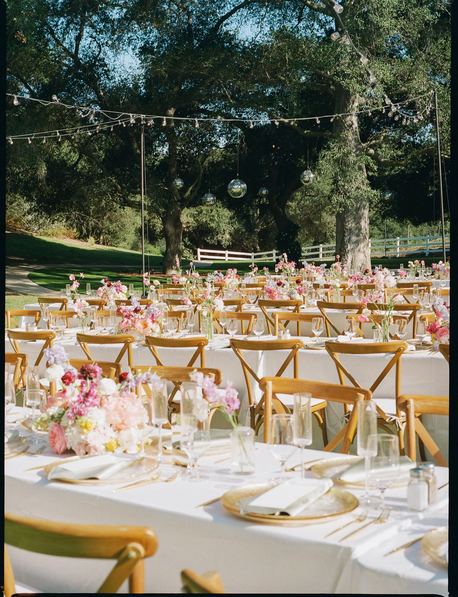 Outdoor wedding reception setup with round tables covered in white tablecloths, decorated with pink and white floral centerpieces, set with glassware, plates, and gold utensils. Wooden chairs surround the tables. String lights and reflective disco balls are hung from trees in a lush green setting with a white fence in the background. The scene is brightly lit, indicating daytime.