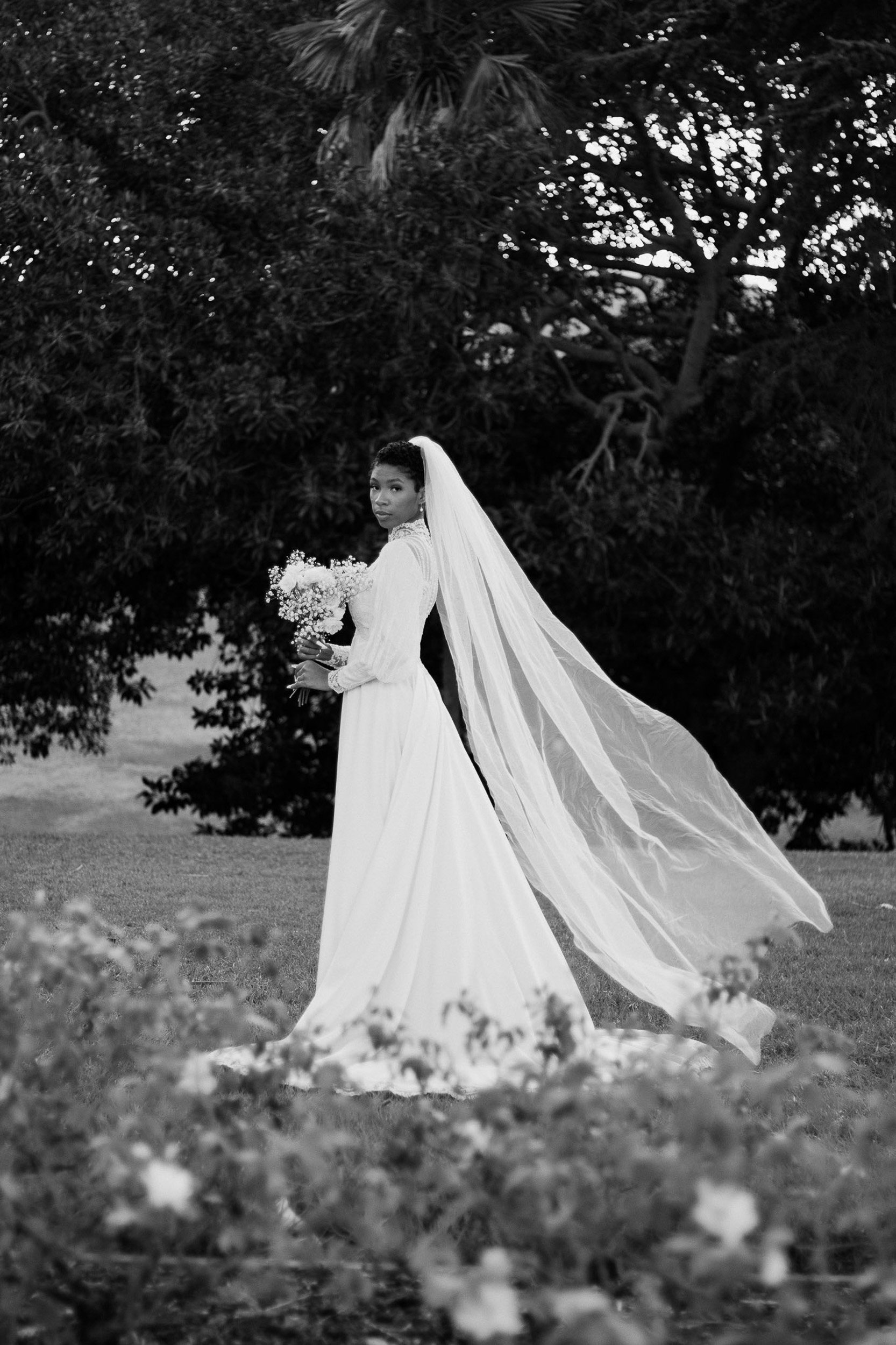 A bride with short curly hair wearing a long-sleeved wedding dress and veil, holding a bouquet of flowers, standing outdoors in front of a large tree.