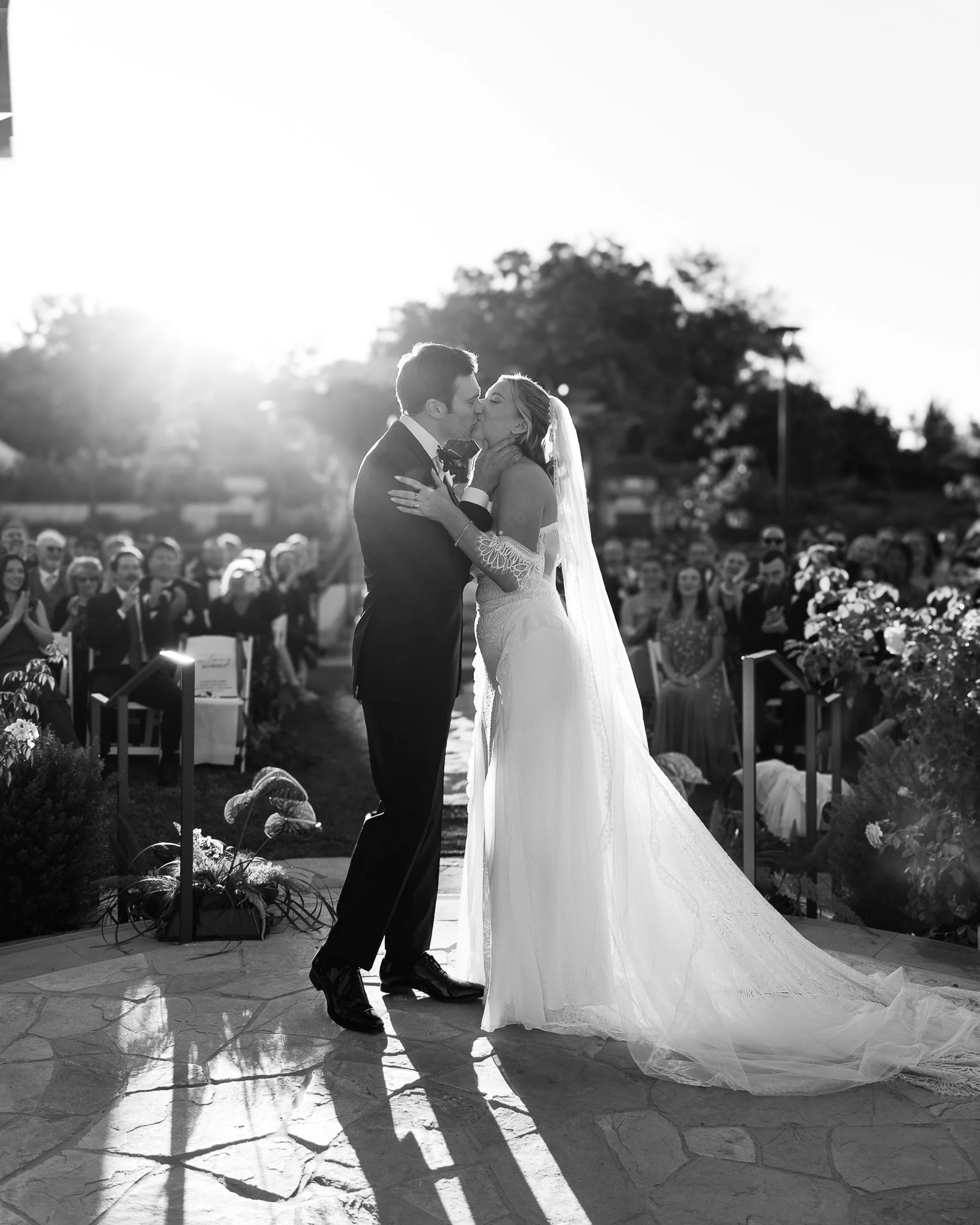 Black and white wedding ceremony photograph of a bride and groom sharing their first kiss at sunset.