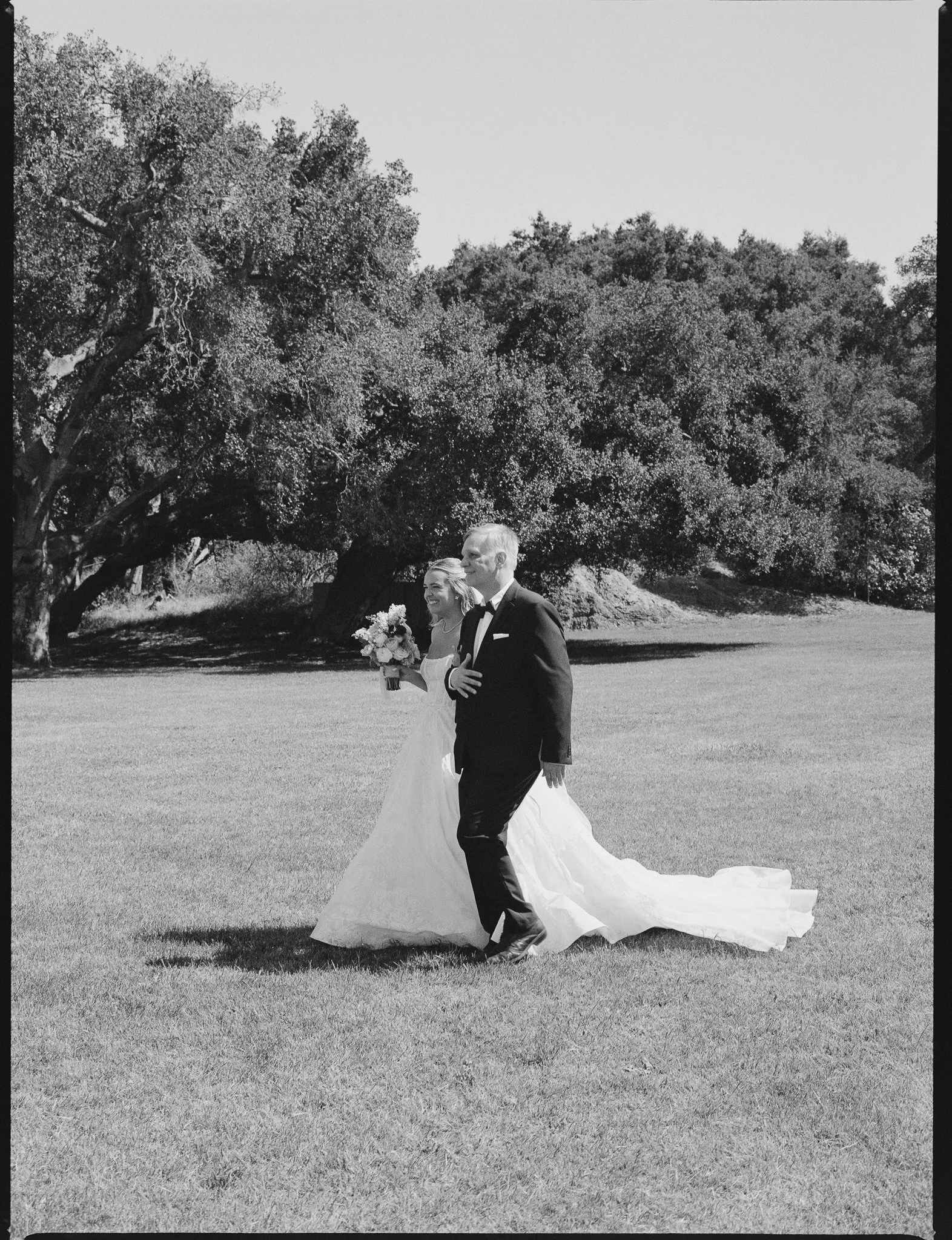 A black and white photo of a wedding couple walking outdoors on grass with large trees in the background. The bride is holding a bouquet and wearing a long white gown, while the groom is dressed in a black tuxedo.