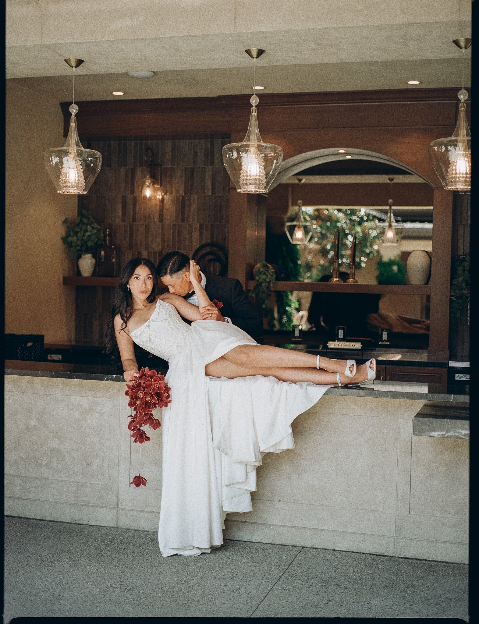 A woman in a white dress is sitting on a bar counter with a man in a black suit kneeling beside her, surrounded by decorated interior space with pendant lights and greenery, and a large mirror in the background.