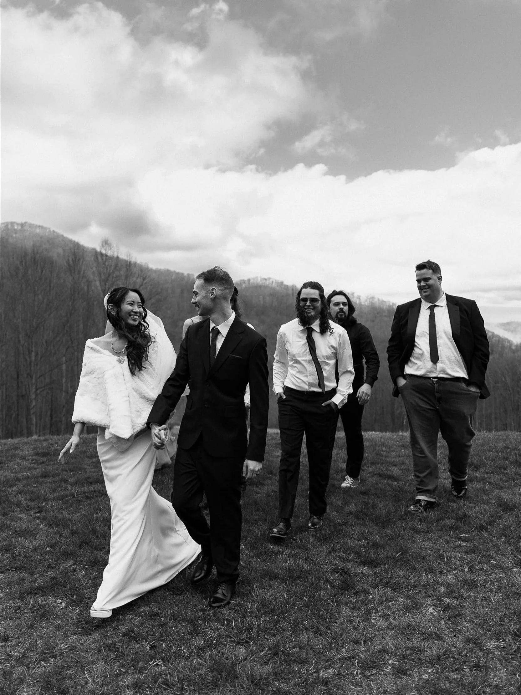 A group of six people, including a bride in a wedding gown and others in formal attire, walking outdoors on grass with a mountain and cloudy sky in the background, smiling and holding hands.