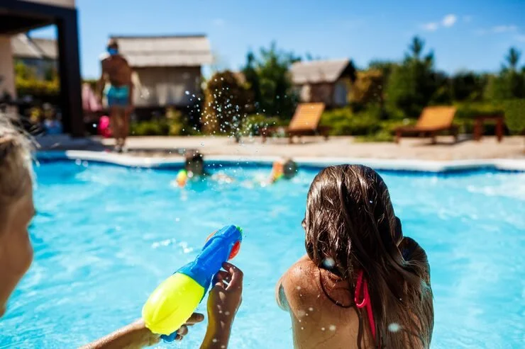 Children playing with water guns in a backyard pool, sunny day with lounge chairs and houses in the background.