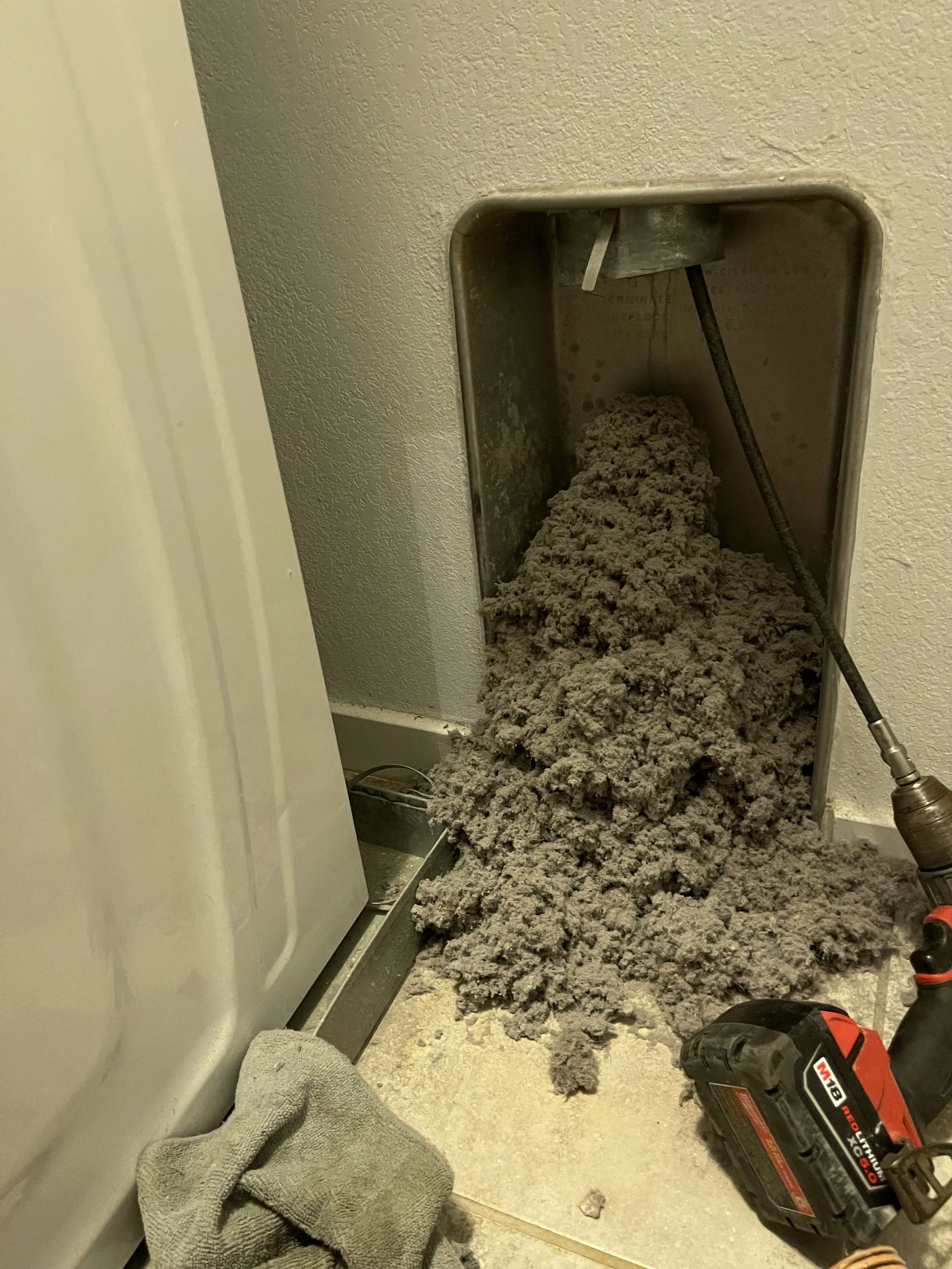 Clogged dryer vent with lint inside the vent opening, a cordless drill on the floor nearby, and laundry towels on the ground.
