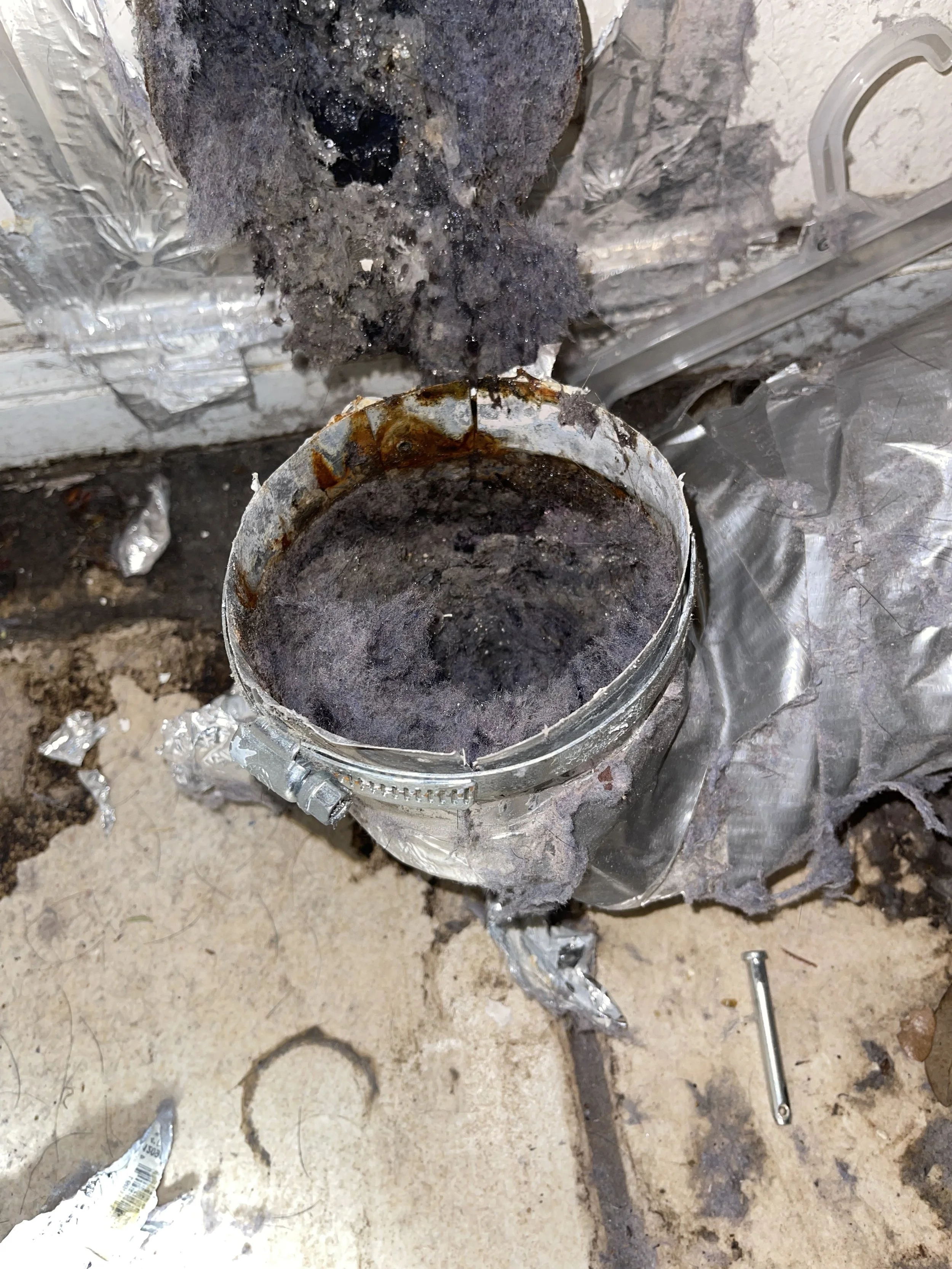 Close-up view of a metal pipe with rust and soot inside, surrounded by damaged foil insulation, on a dirty concrete floor with assorted tools and debris nearby.