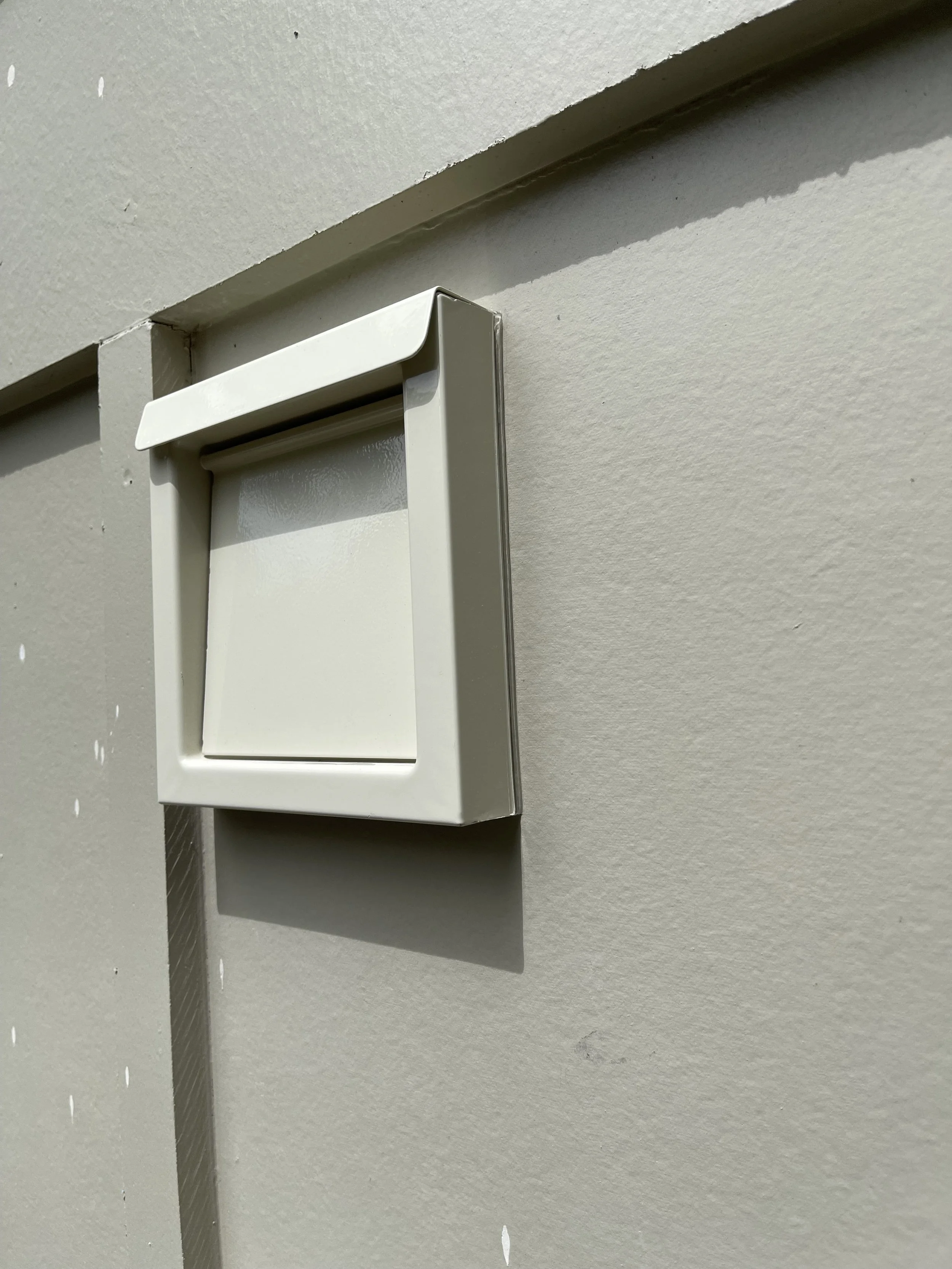 Close-up of a white wall-mounted mailbox with a drop-shaped opening, situated on a beige wall with a shadow cast from the mailbox.