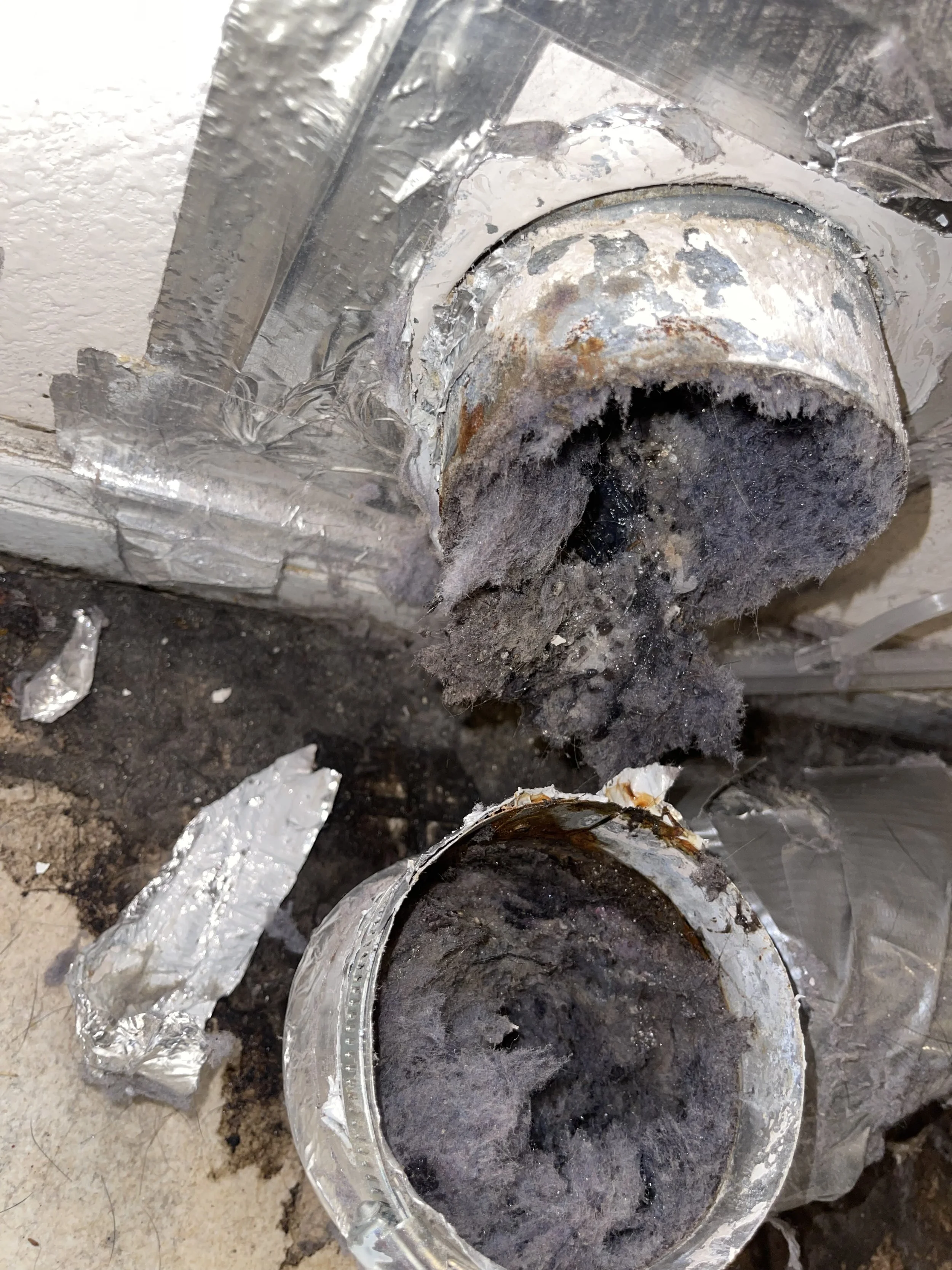 Close-up of a damaged and rusted pipe with some broken aluminum foil around it, on a dirty ground surface.