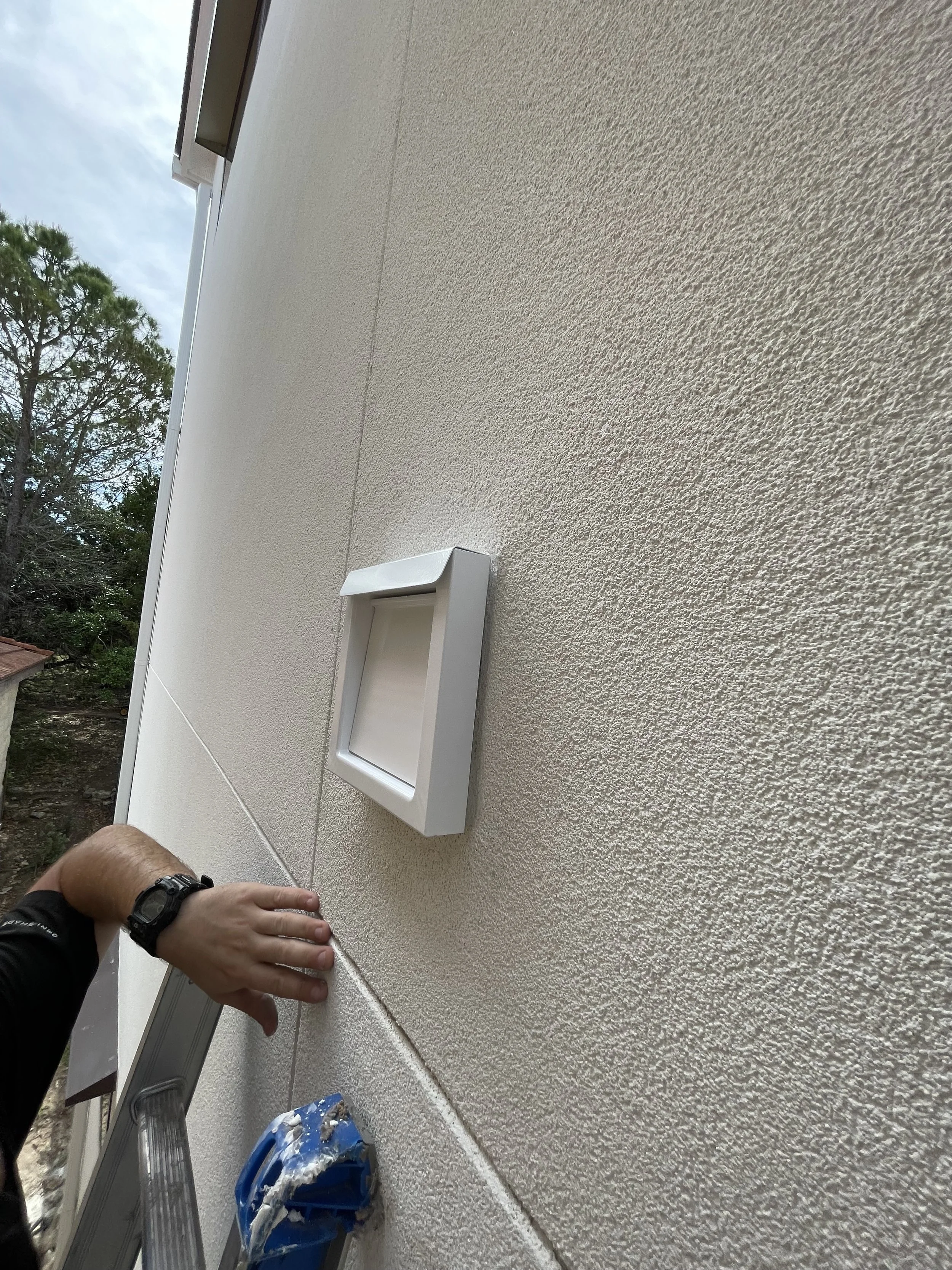 Person installing a white outdoor electrical or utility box on a textured beige exterior wall, with a ladder and a paint sprayer visible in the scene.