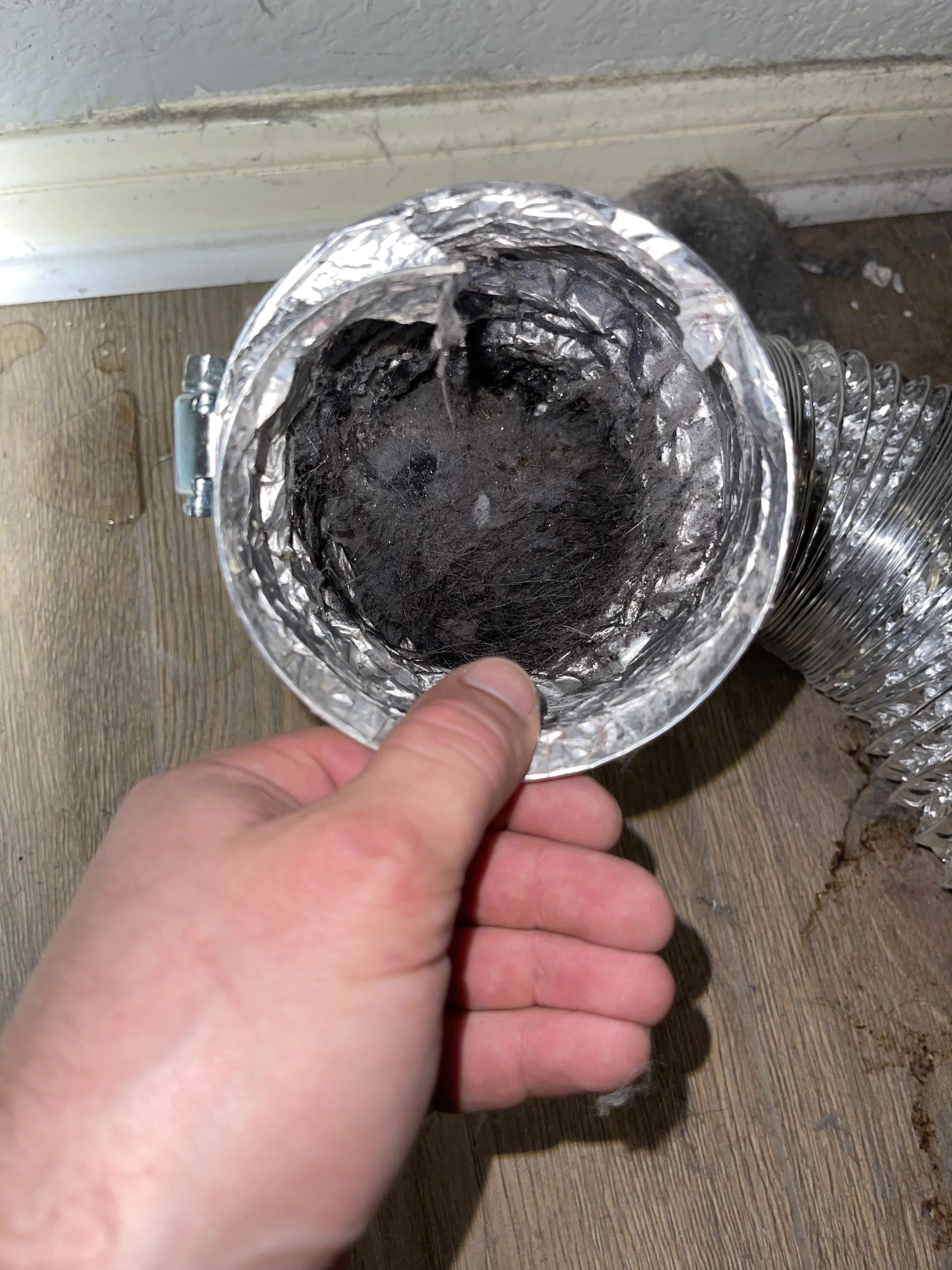 Close-up of a person holding a dusty, reflective metal duct with foil insulation inside, with more duct segments stacked on a wooden surface nearby.