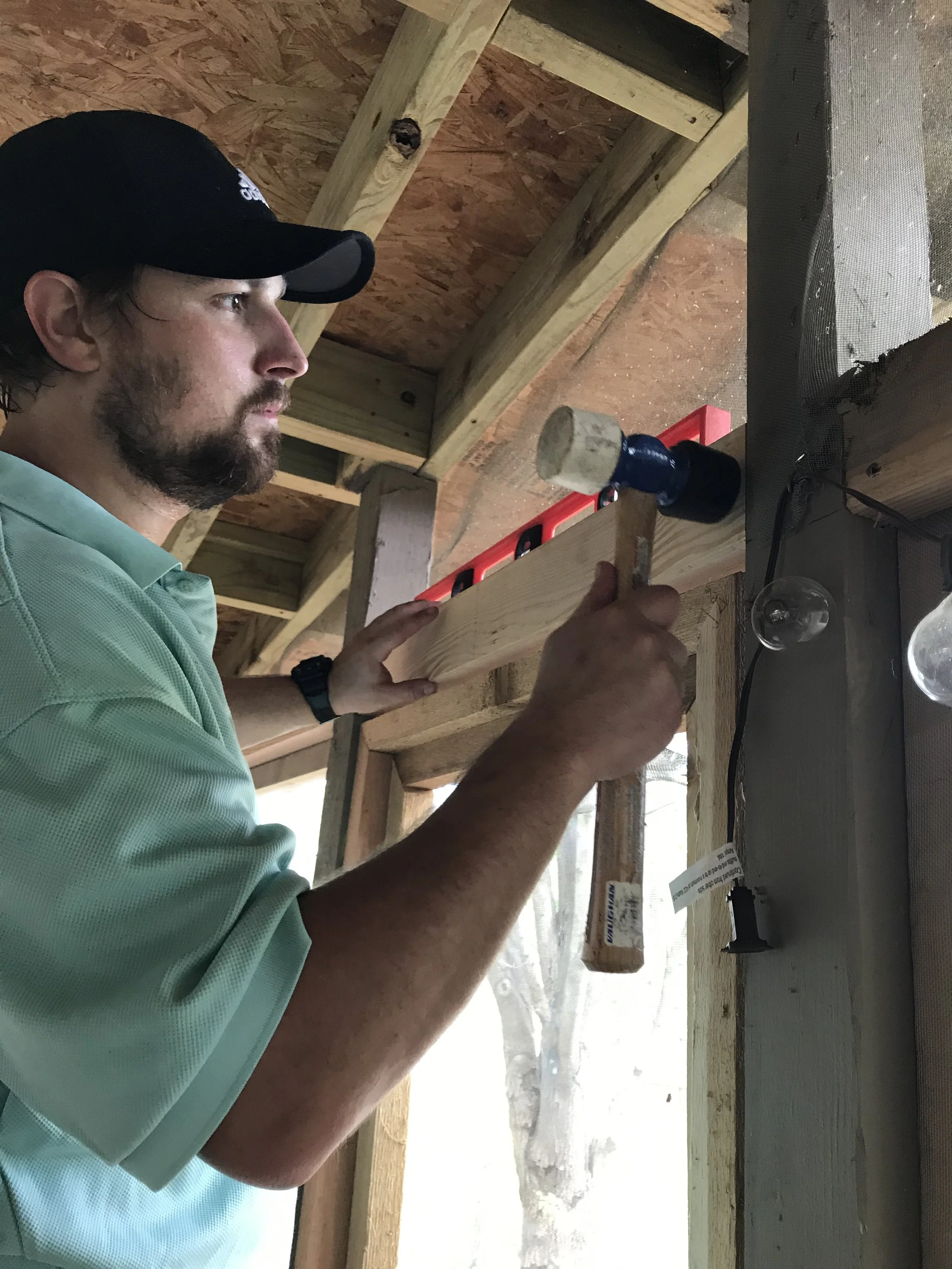 A man in a black cap and mint green shirt using a hammer on a piece of wood at a construction site with exposed wooden framing and plywood walls.