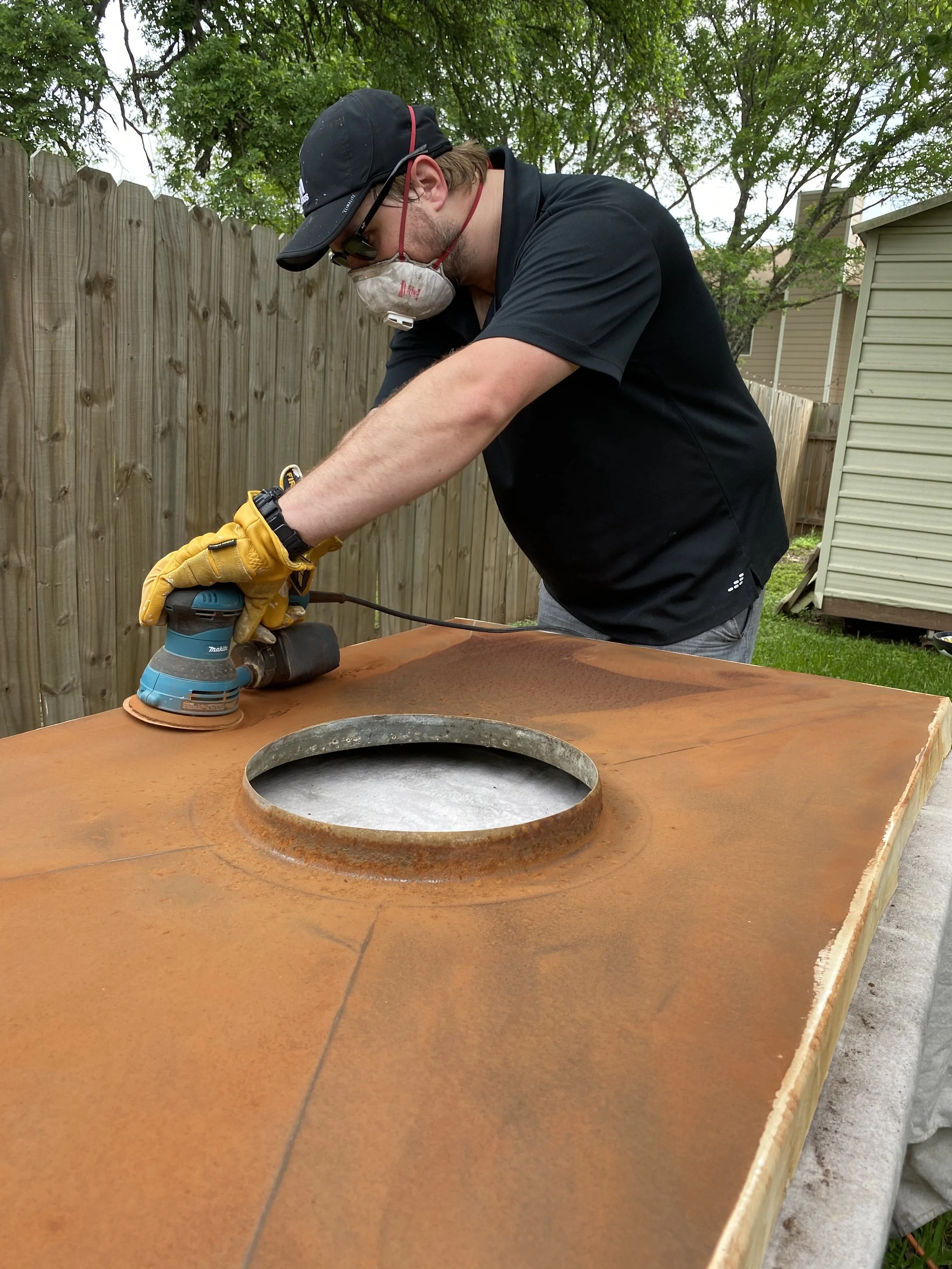A man wearing a black cap, protective mask, sunglasses, and yellow gloves is using a handheld power sander to sand the surface of a wooden table outdoors. There is a wooden fence and a house in the background.
