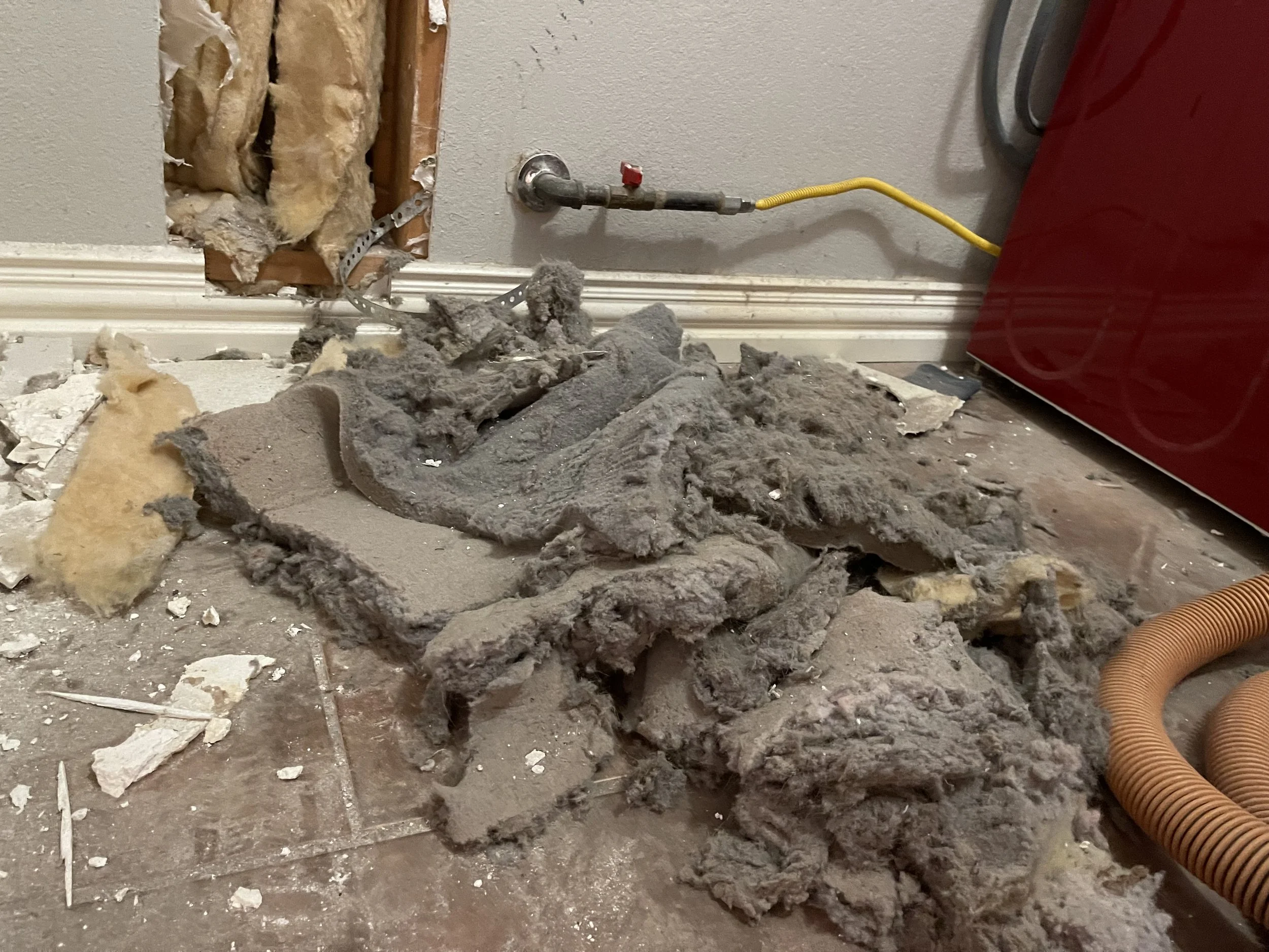 Hole in the wall with exposed insulation, debris, and pipes including a yellow electrical wire and an orange flexible vent pipe.