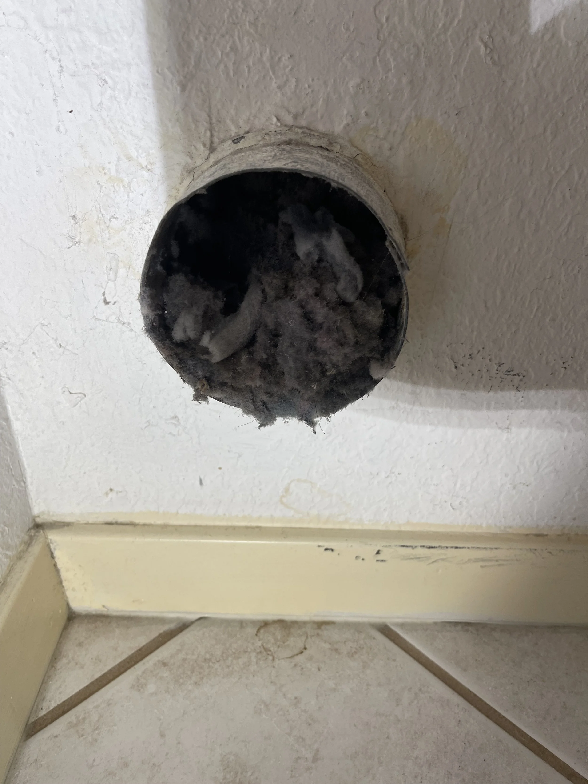 An unfinished or damaged bathroom wall with a round pipe opening, surrounded by dirt and debris, near a corner with tile and baseboard.