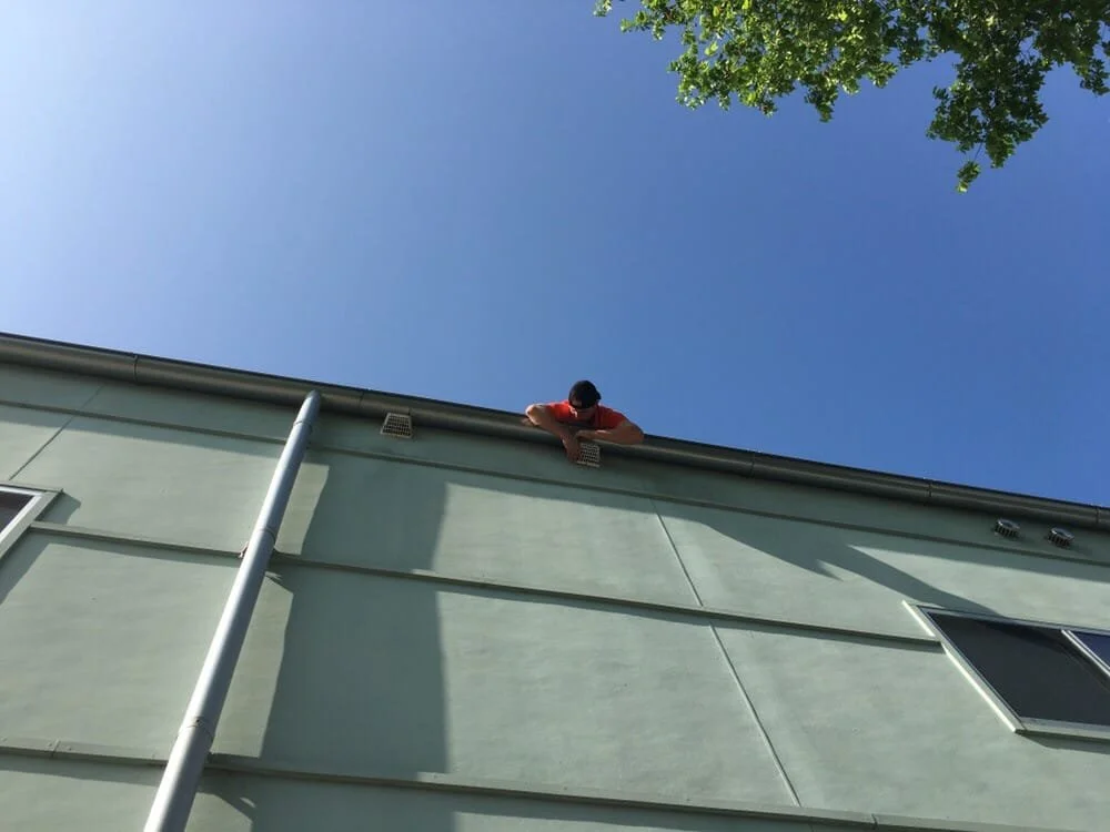 A person wearing an orange shirt and dark hat leaning over the edge of a house roof, looking down, with a clear blue sky and some green tree branches in the background.