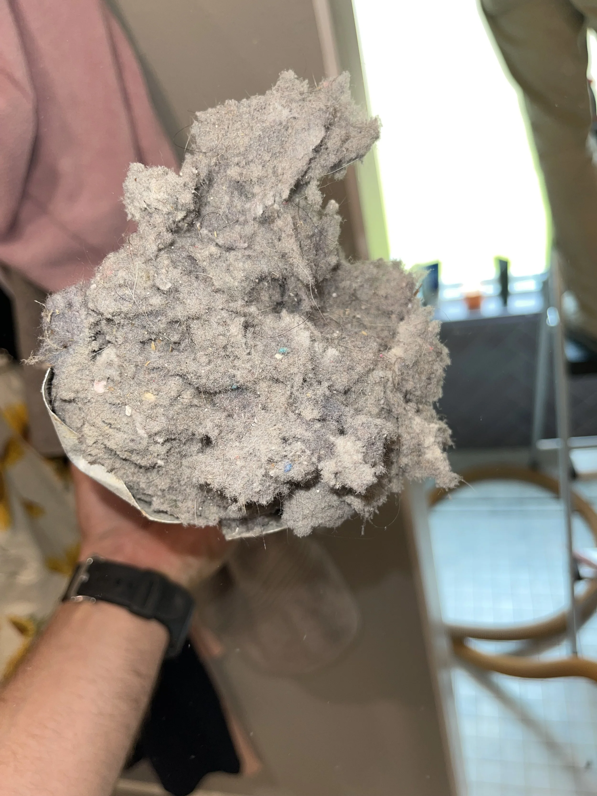 A hand holding a large clump of gray dust or debris, with visible fibers and small particles on its surface, near a window and wooden chairs.