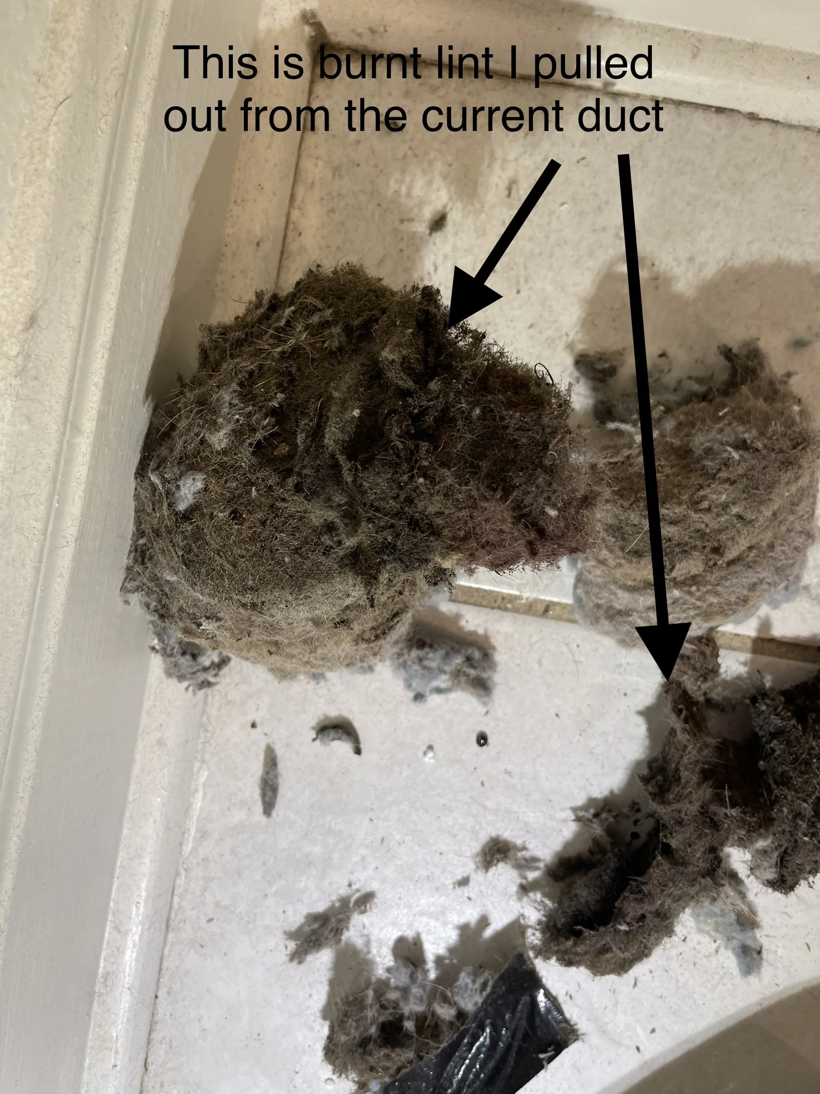 Close-up of a burnt lint ball on a white surface with a note pointing to it, saying 'This is burnt lint I pulled out from the current duct.' Burnt lint and debris scattered around.
