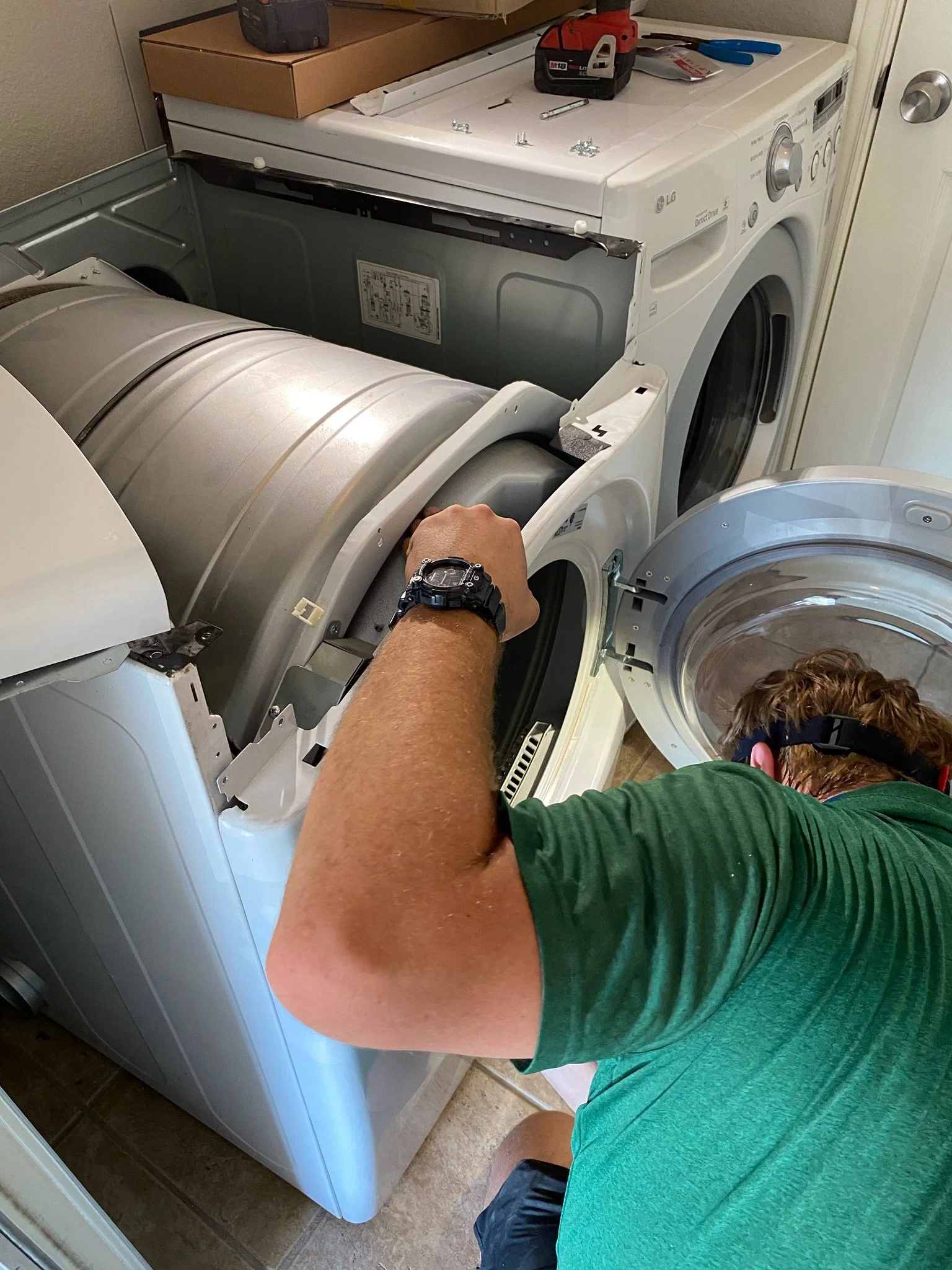 A person wearing a green shirt and a black watch is fixing a top-loading washing machine, with parts and tools on top of the machine and a person in the background.