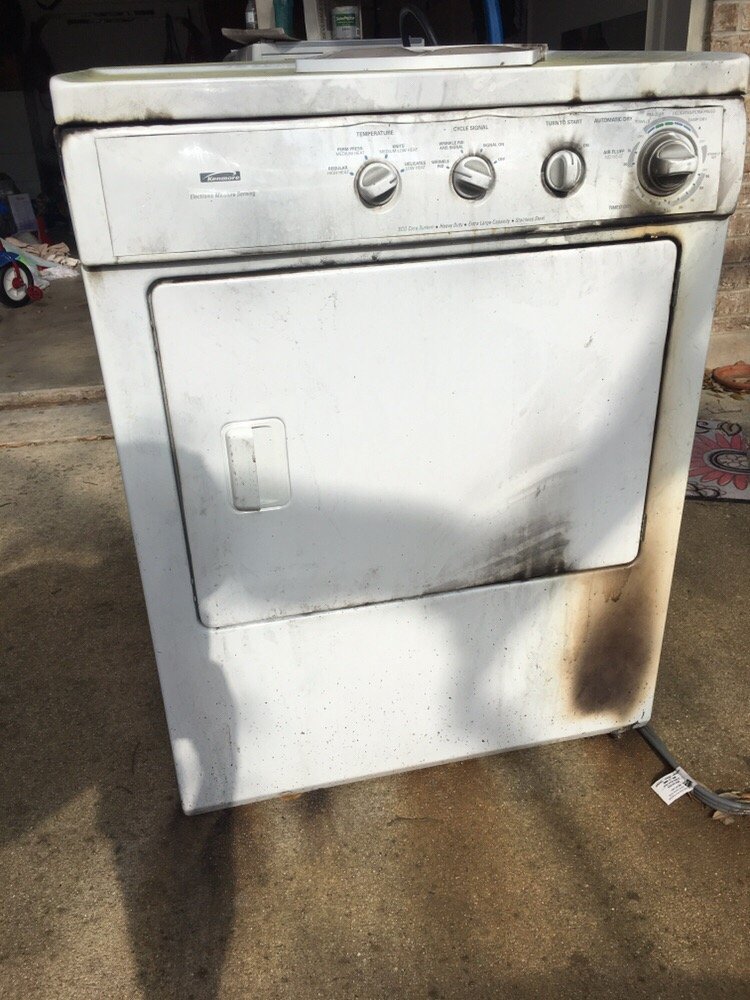 A white, old, dirty clothes dryer with rust and stains on the exterior, situated outdoors on a concrete surface, with a power cord on the ground nearby.