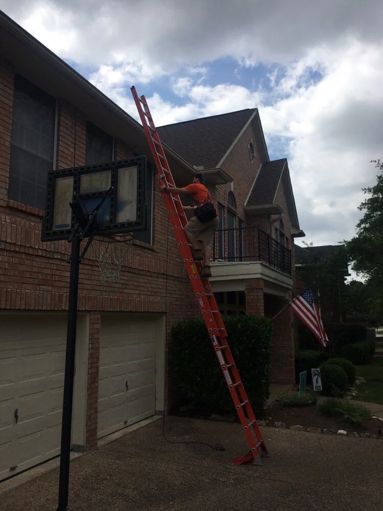 A person standing on a tall, orange extension ladder cleaning a second-story window of a brick house.