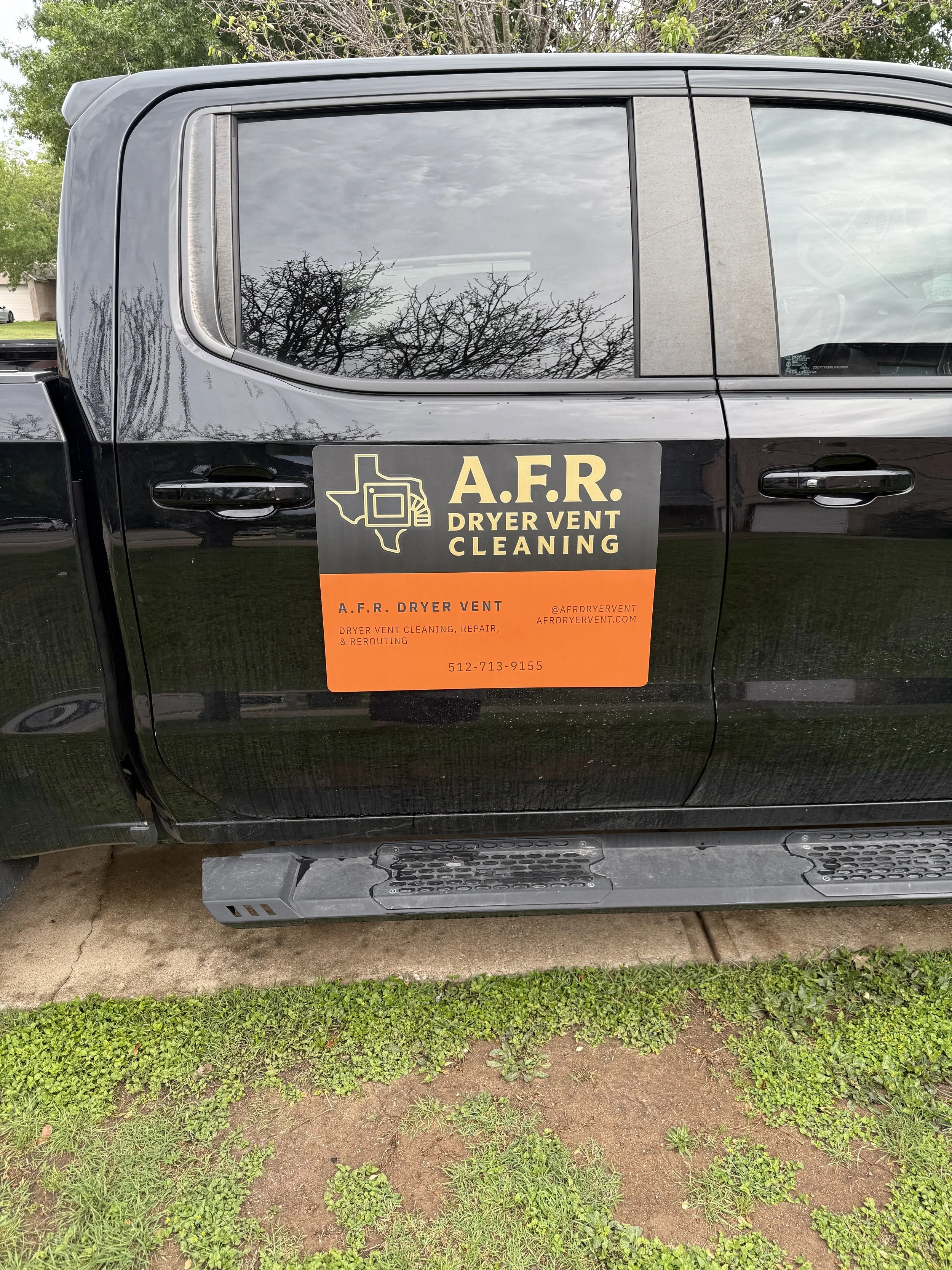 The image shows the side of a black pickup truck parked on a grassy and dirt surface. The truck has a large color sign on its side advertising A.F.R. Dryer Vent Cleaning with a phone number and website. The background includes trees and a residential