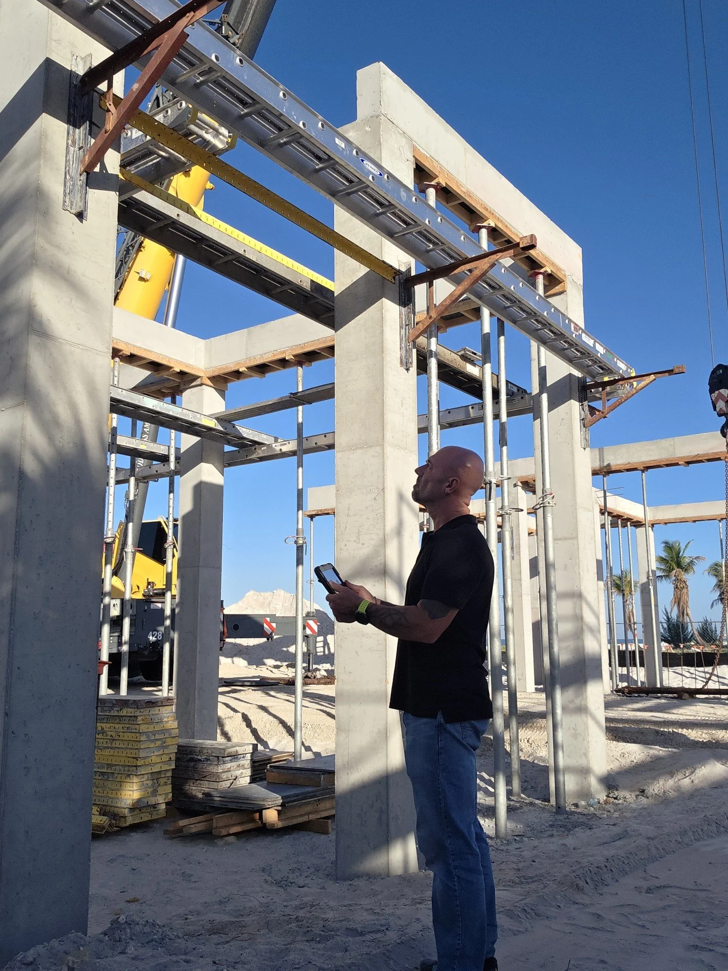 A man standing on a construction site looking at the framework of a building under construction, with scaffolding, concrete, and a clear blue sky.