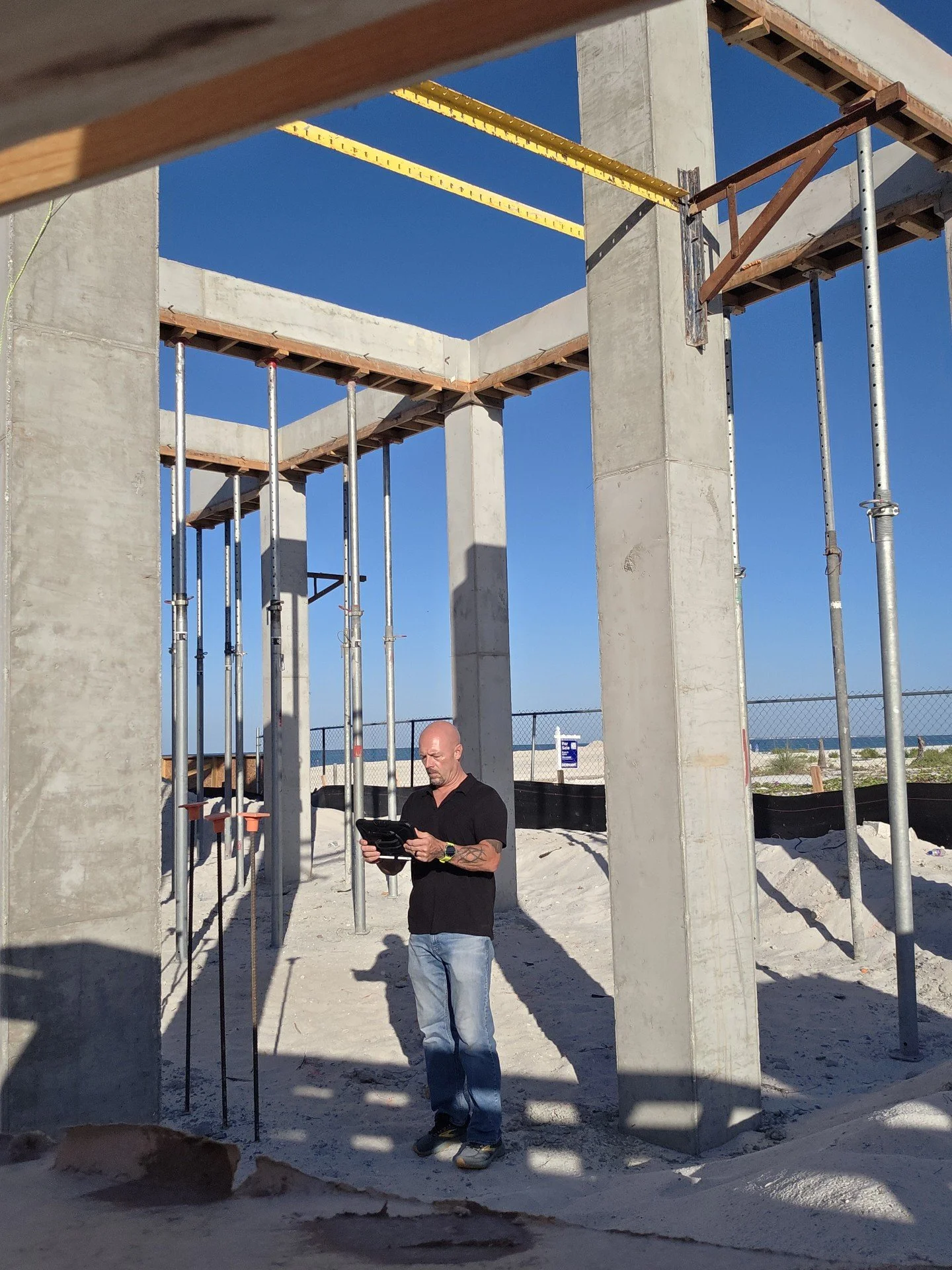 A man standing in a construction site beneath a concrete structure, holding and looking at a tablet or device, with scaffolding around him and a clear blue sky overhead.