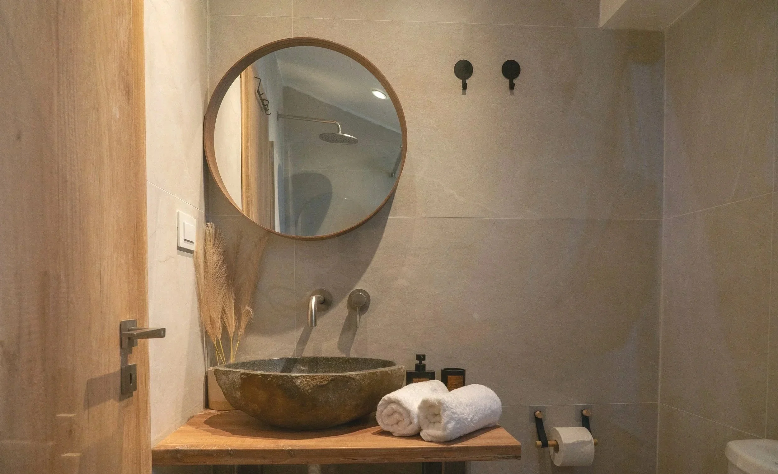 A modern bathroom with a wooden vanity holding a stone basin sink, with rolled white towels and soap containers. There is a round mirror on the wall reflecting a shower area with a rain showerhead, a wall-mounted faucet, and two empty hooks. The bathroom has beige tiled walls and a minimalist decor.