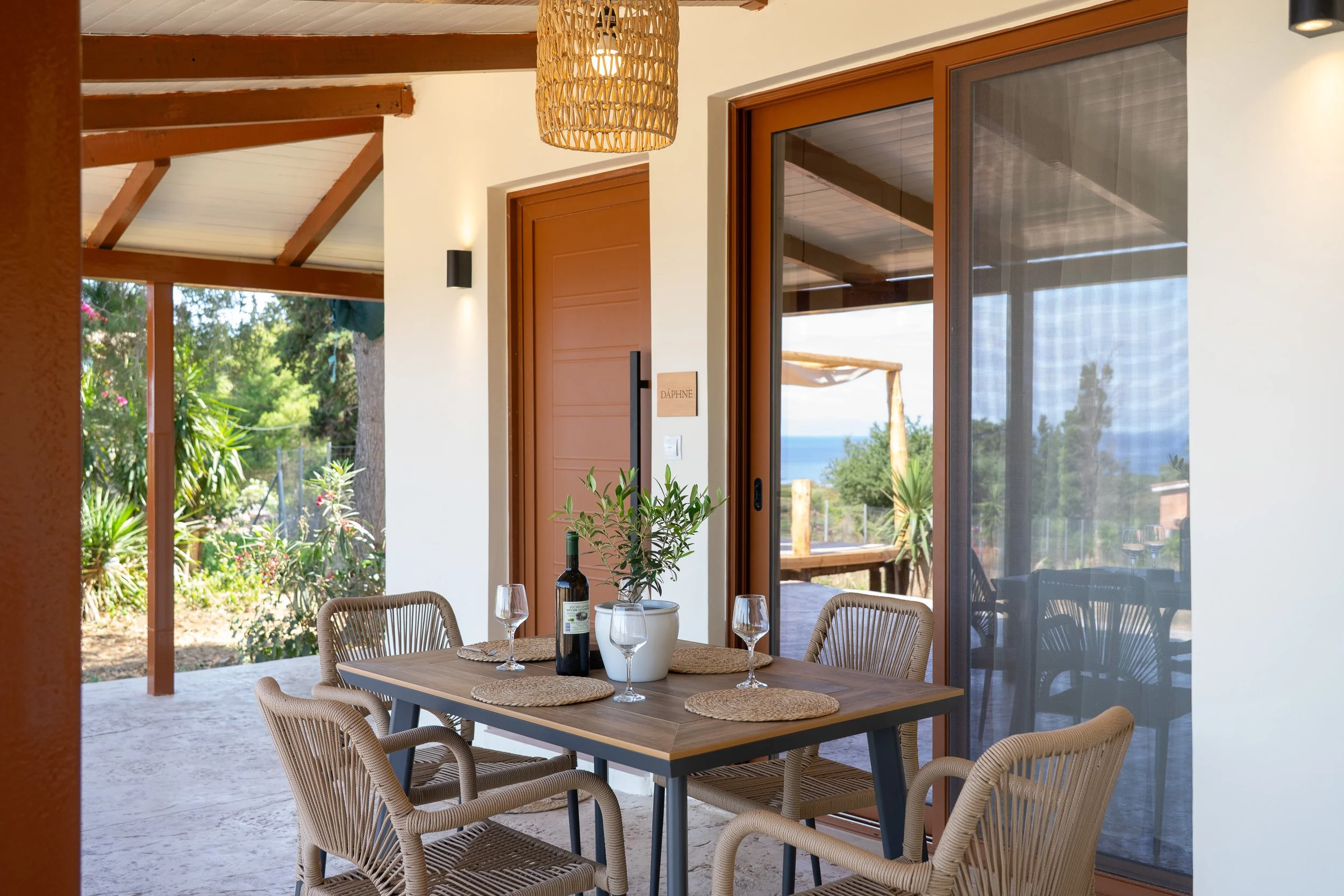 Outdoor patio dining area with wooden table, six wicker chairs, a wine bottle, four glasses, a potted olive tree, and a view of the ocean in the background.