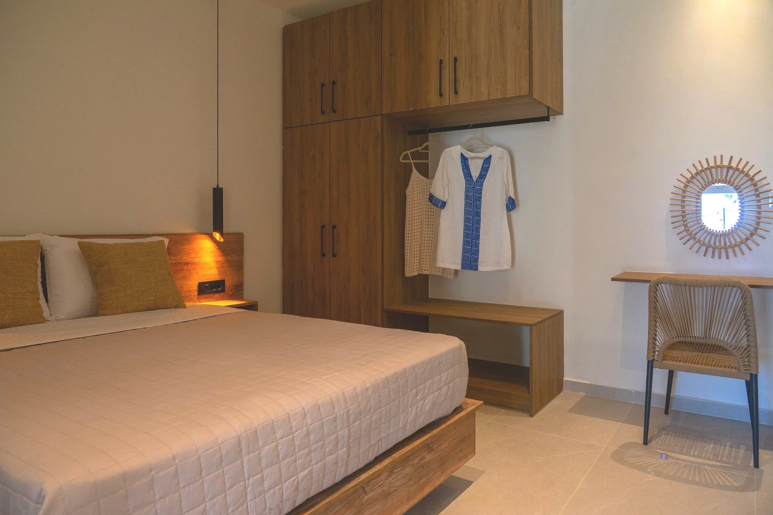 Bedroom with a bed, wooden headboard, yellow pillows, wardrobe, hanging clothes, wooden bench, mirror, and chair.