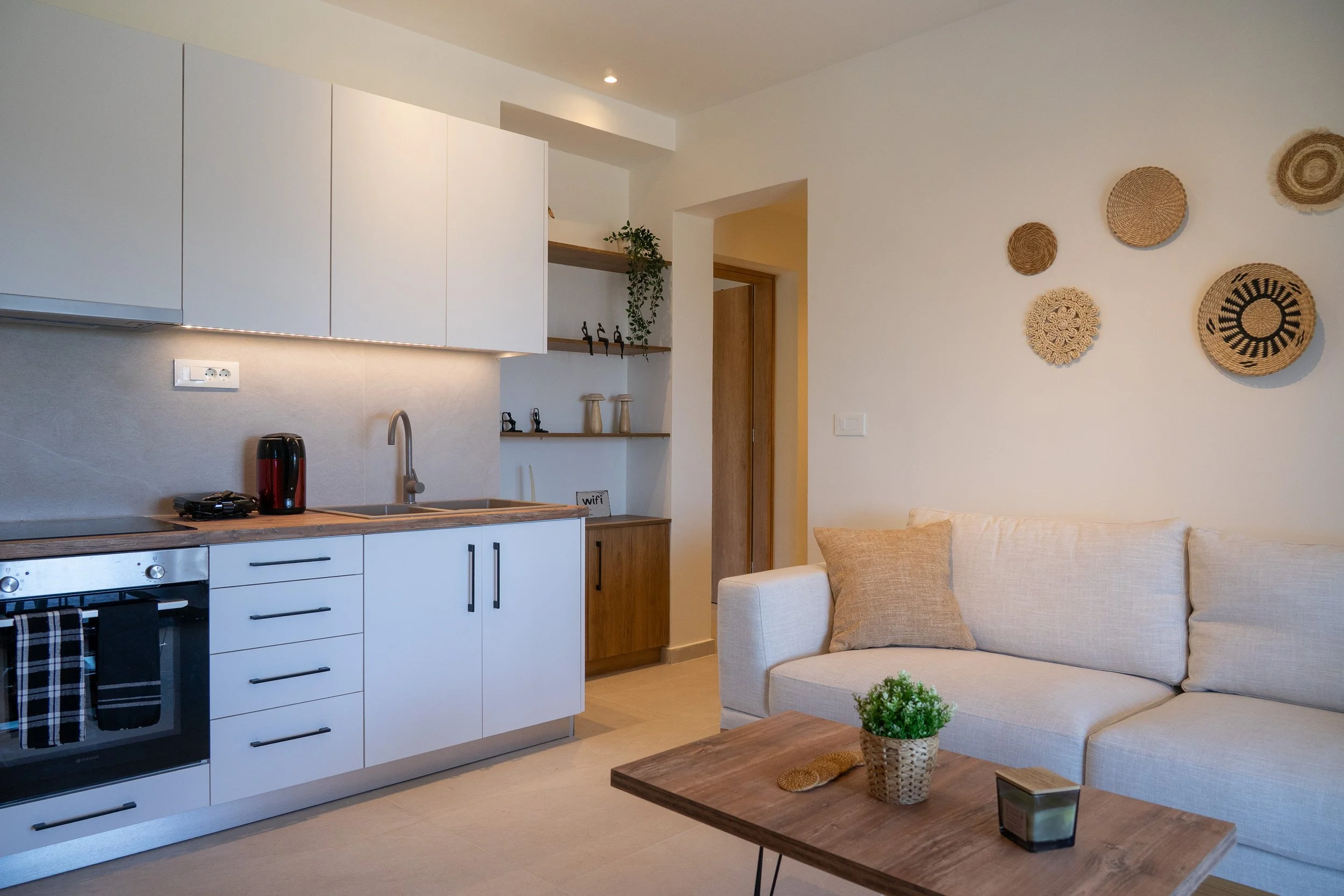 Living room with a white couch, decorative wall hangings, a wooden coffee table with a small basket plant, and an open kitchen area with white cabinets, a wooden countertop, a stove, and a kettle.