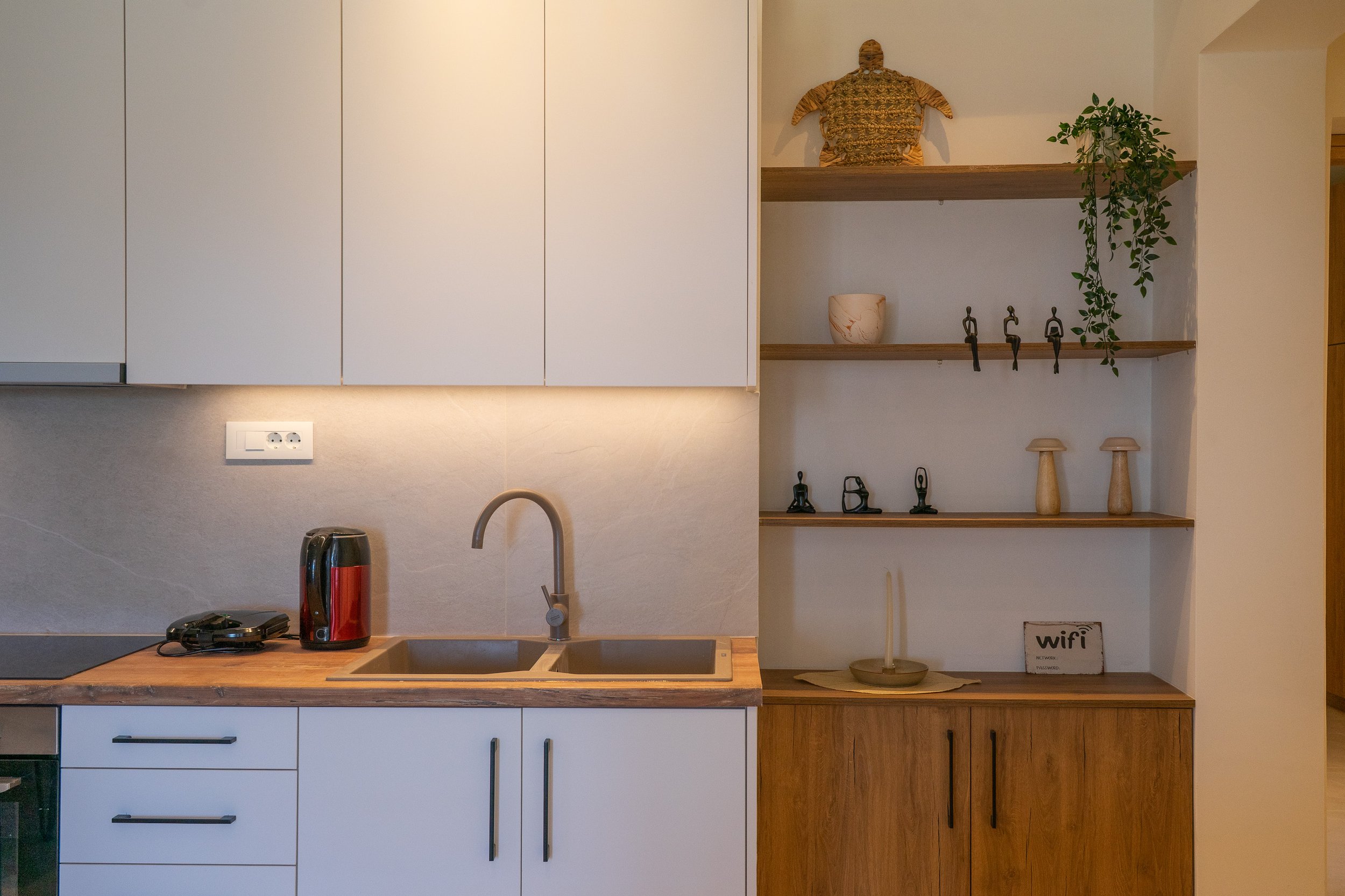 Kitchen with white cabinets, a wooden countertop, a black and red electric kettle, a black stovetop, a remote control, a double sink, a beige backsplash with an electrical outlet, and open wooden shelves with decorative items including small sculptur