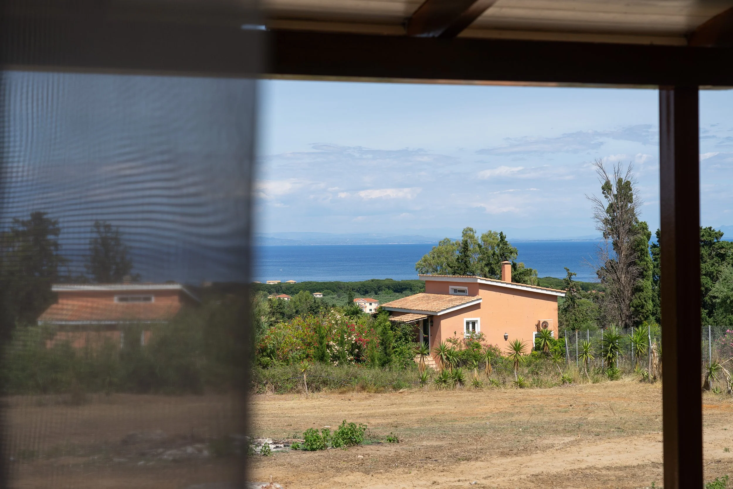 View of a house with terracotta roof and peach-colored walls, surrounded by lush greenery and palm trees, overlooking a large body of water under a partly cloudy sky.