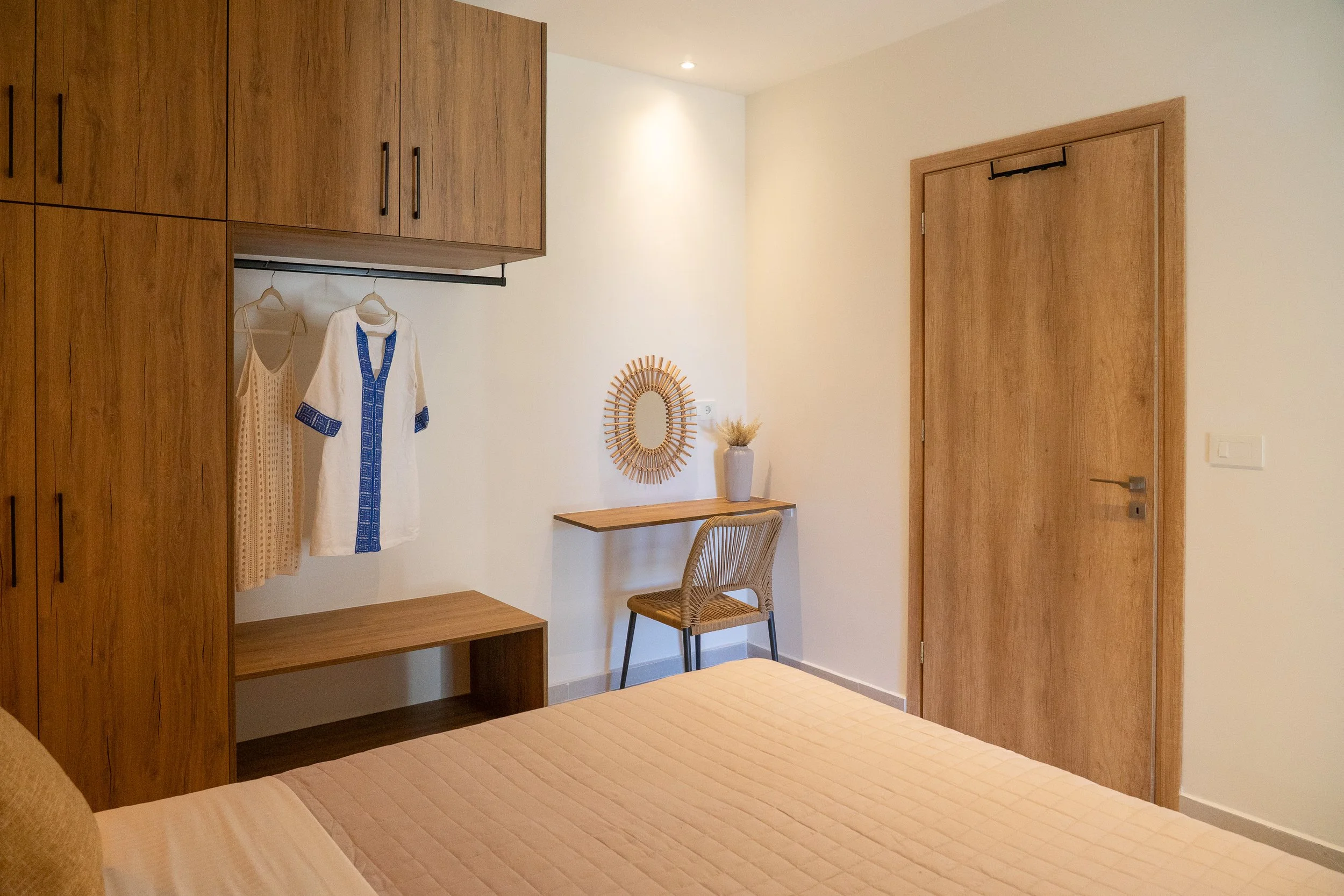 A bedroom with a wooden wardrobe, a small desk with a vase and dried flowers, a wicker chair, a mirror on the wall, and a bed with a beige quilt.