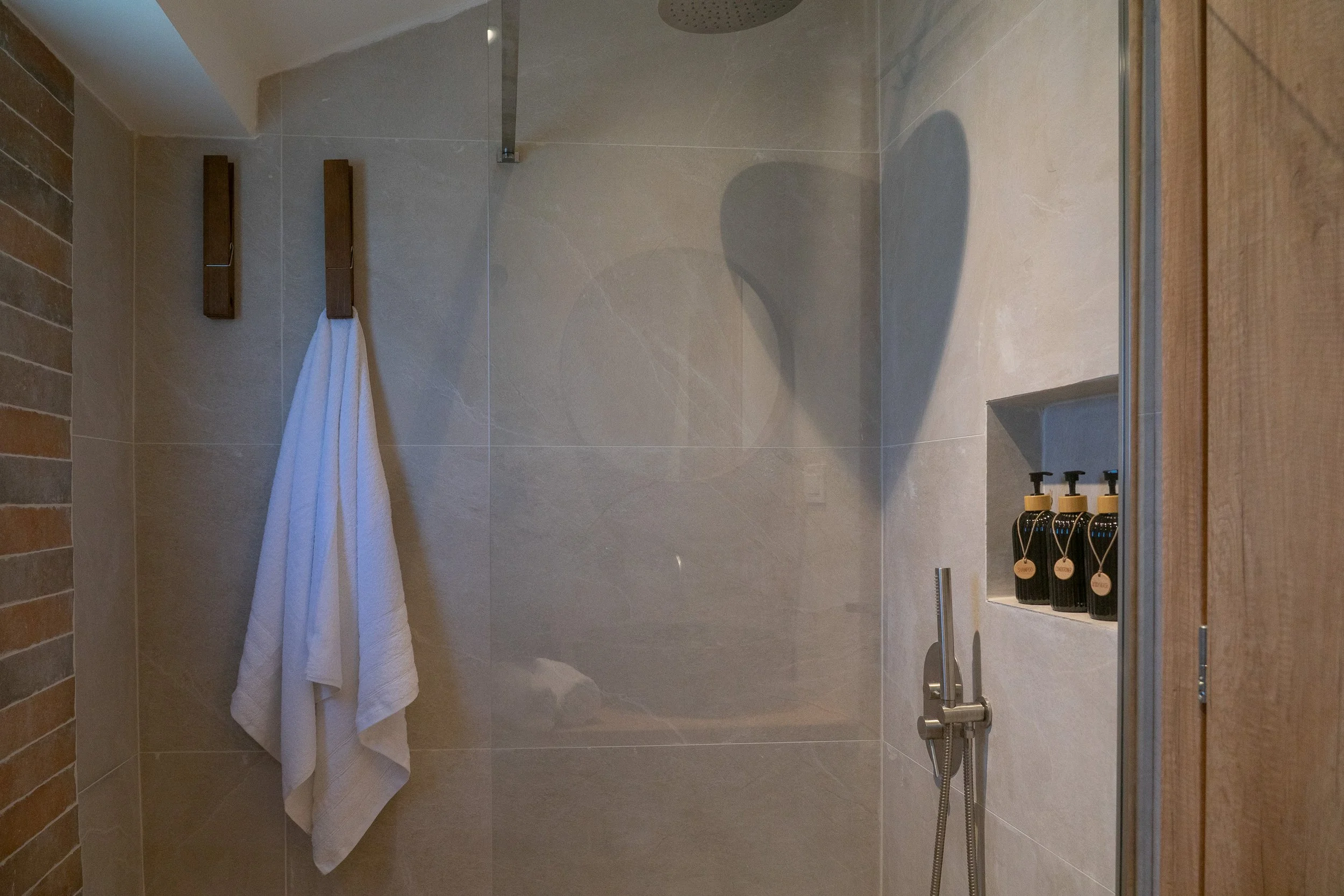 A modern shower area with beige tiled walls, a wooden towel rack holding a white towel, and a built-in niche with three black bottles of bath products. There is a shower head mounted on the ceiling and a handheld shower with a hose.
