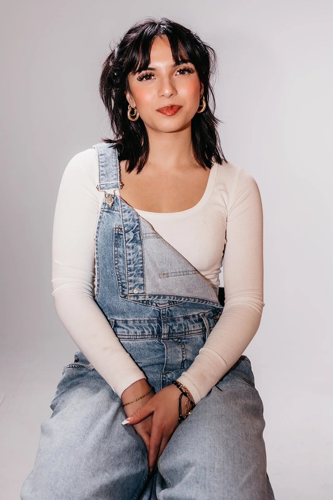 A young woman with dark hair and gold hoop earrings, wearing a white long-sleeve top and denim overalls, sitting against a plain background.