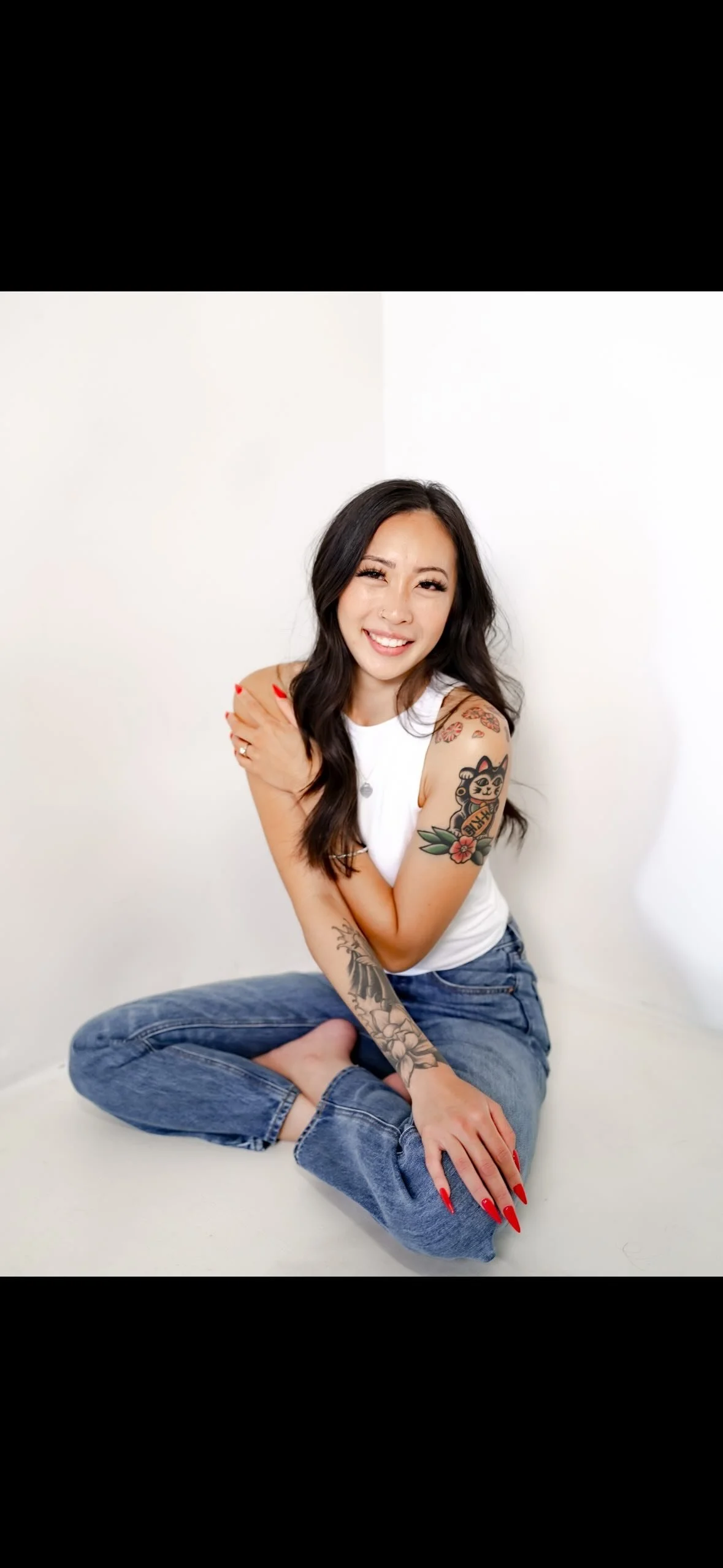 A woman with long black hair and tattoos on her arms sitting cross-legged on a white background, smiling and looking at the camera, wearing a white sleeveless top and blue jeans.