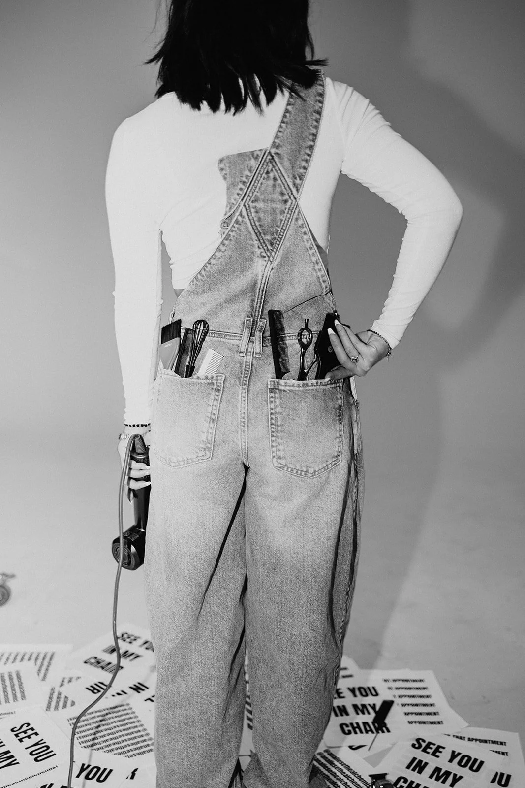 Back view of a person wearing denim overalls with various hair styling tools in the pockets, holding a pair of scissors and hair clippers, with a gray background and text-covered floor.