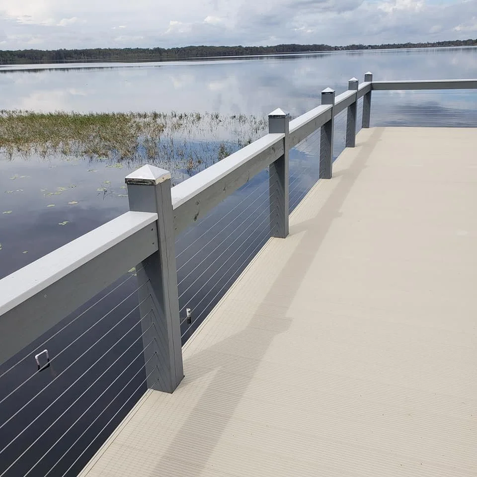 A view of a lake from a wooden pier with a gray railing and beige floor, under a partly cloudy sky.