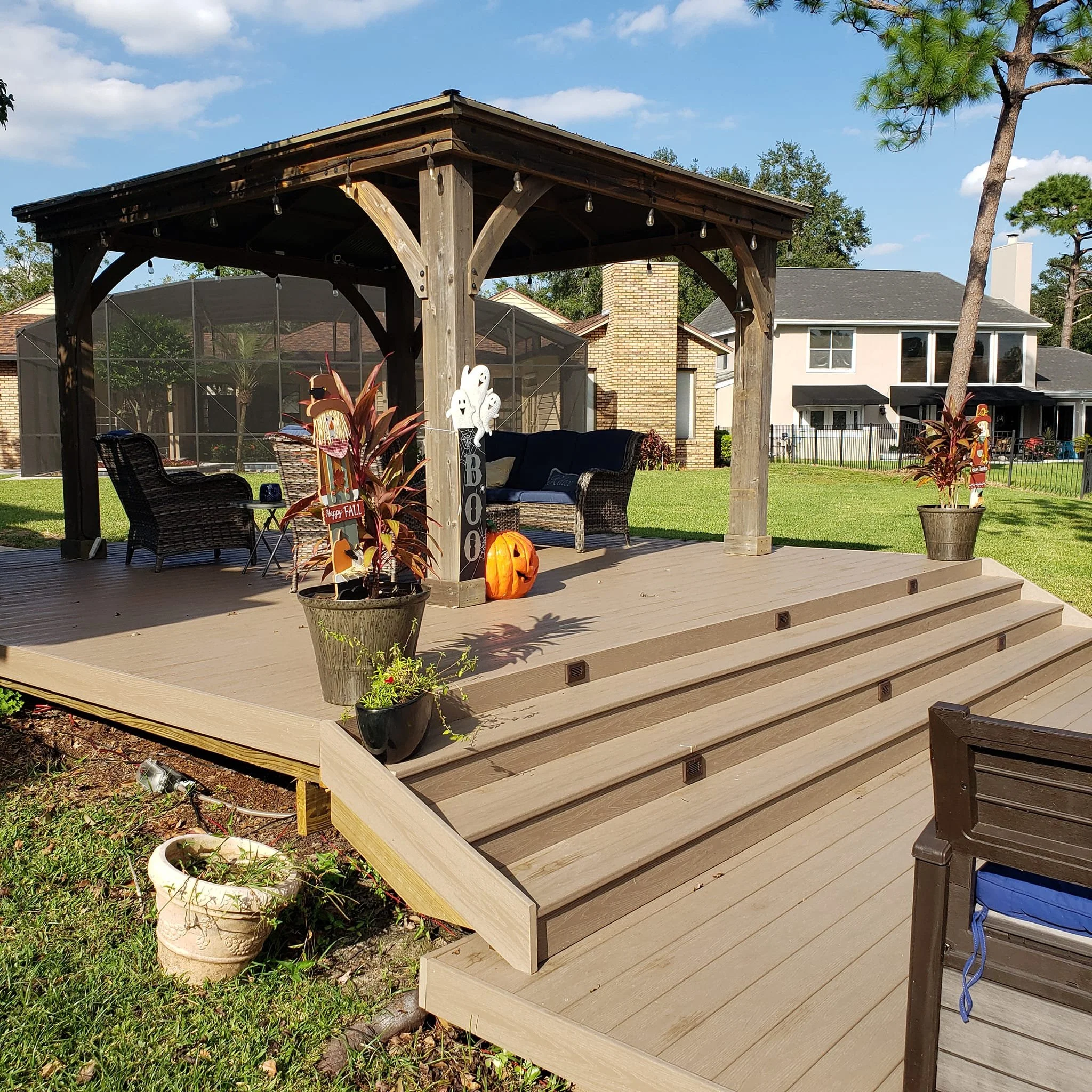 A backyard patio with a wooden pergola, outdoor furniture, potted plants, and nearby houses under a blue sky.