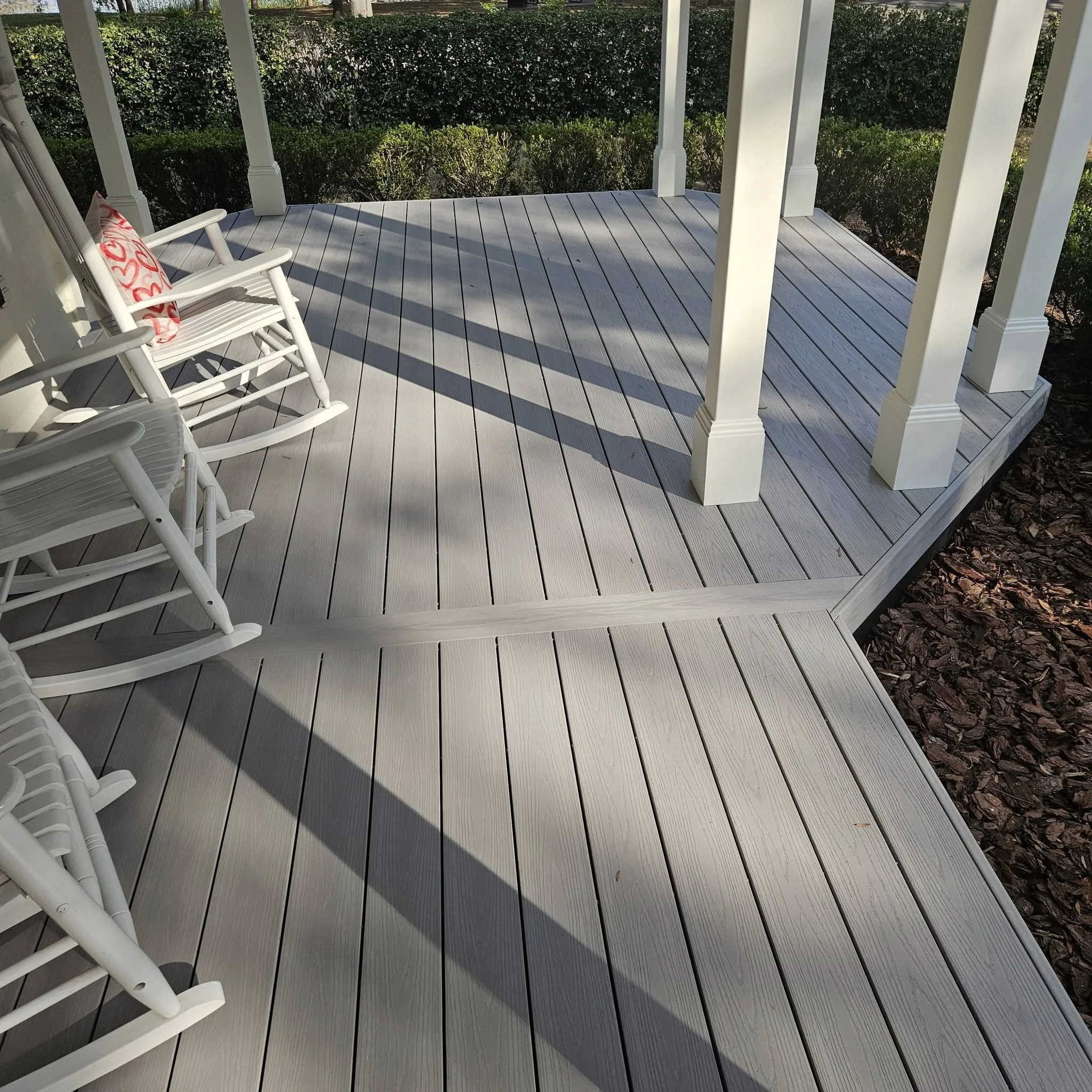 A porch with light gray wooden flooring, white railing and support posts, a white rocking chair with a pink and red patterned pillow, and bushes in the background.